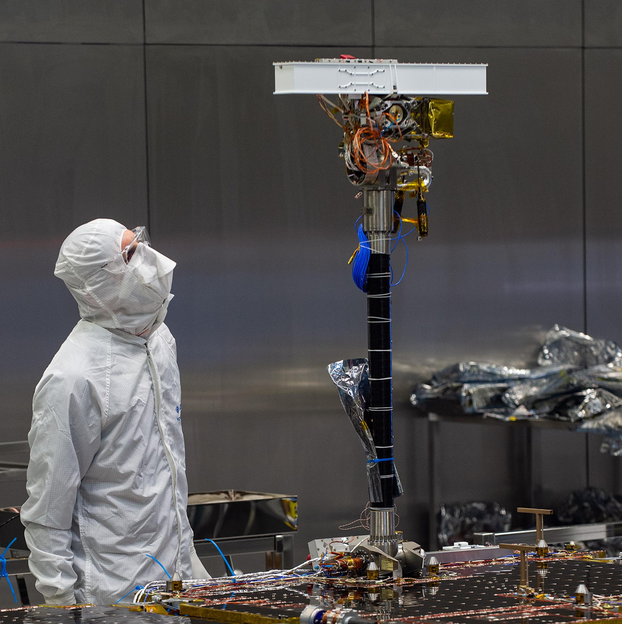 Ein Mann im Schutzanzug mit Gesichtsmaske steht in einem Reinraum und betrachtet den Kamerahals des ExoMars Rover