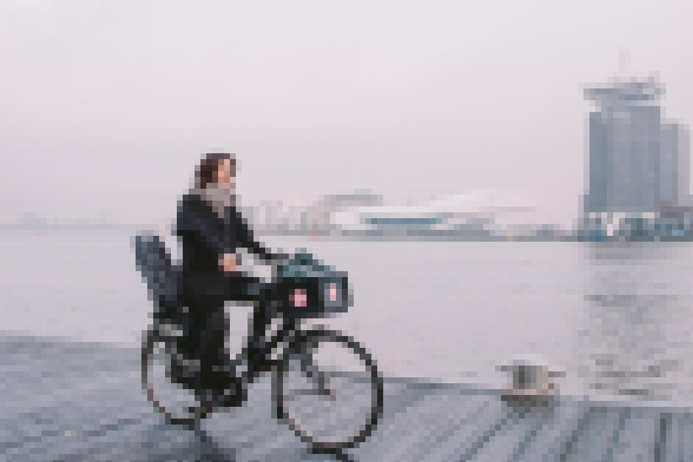 Eine Frau fährt mit Fahrrad auf einem Steg am Wasser. Im Hintergrund sind Hochhäuser zu erkennen.