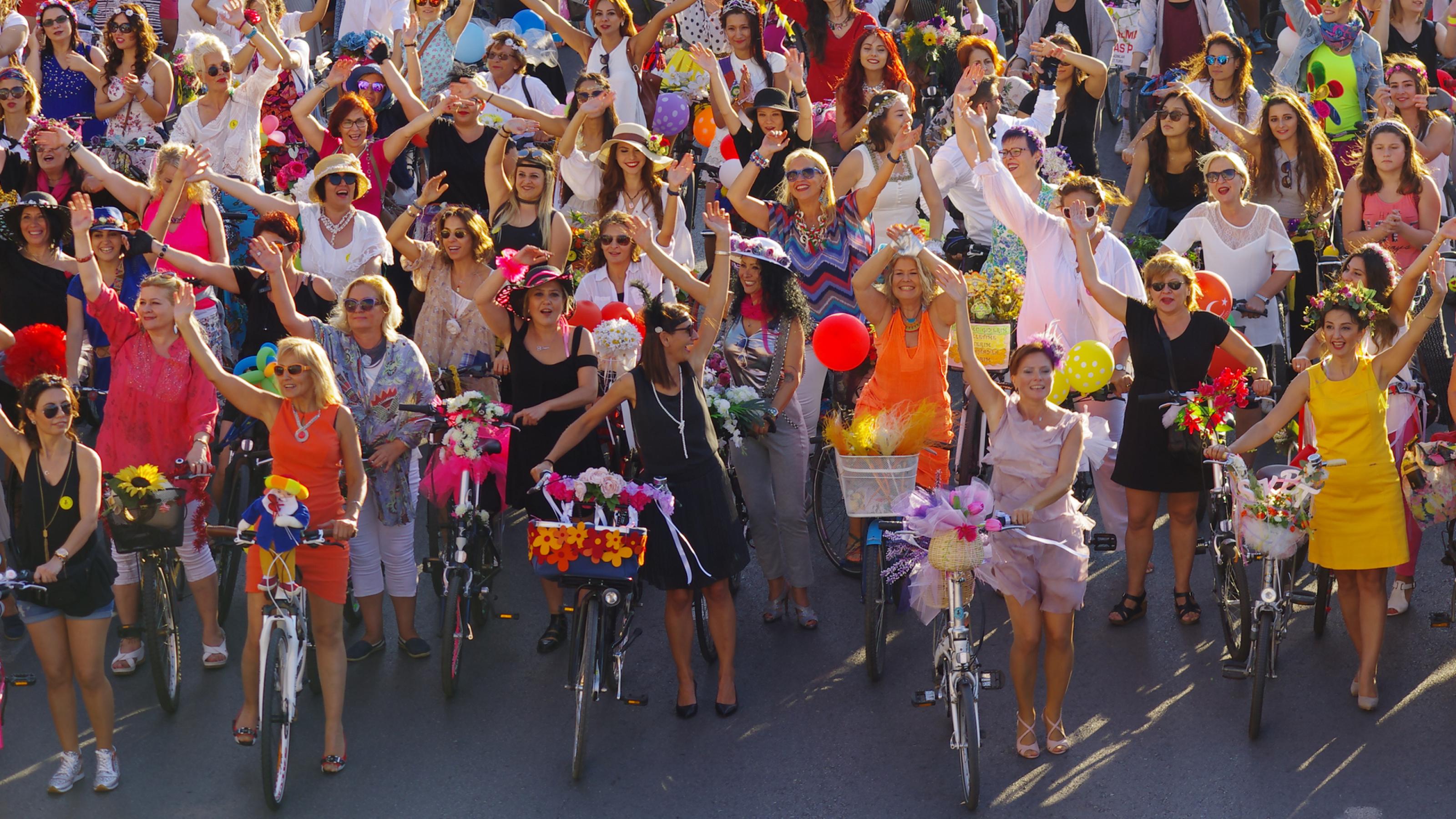 Dutzende von Frauen stehen gut gelaunt mit ihren Rädern in bunten Sommerkleidern nebeneinander und winken in die Kamera. Ihre Körbe haben sie mit Blumen und Federn geschmückt.