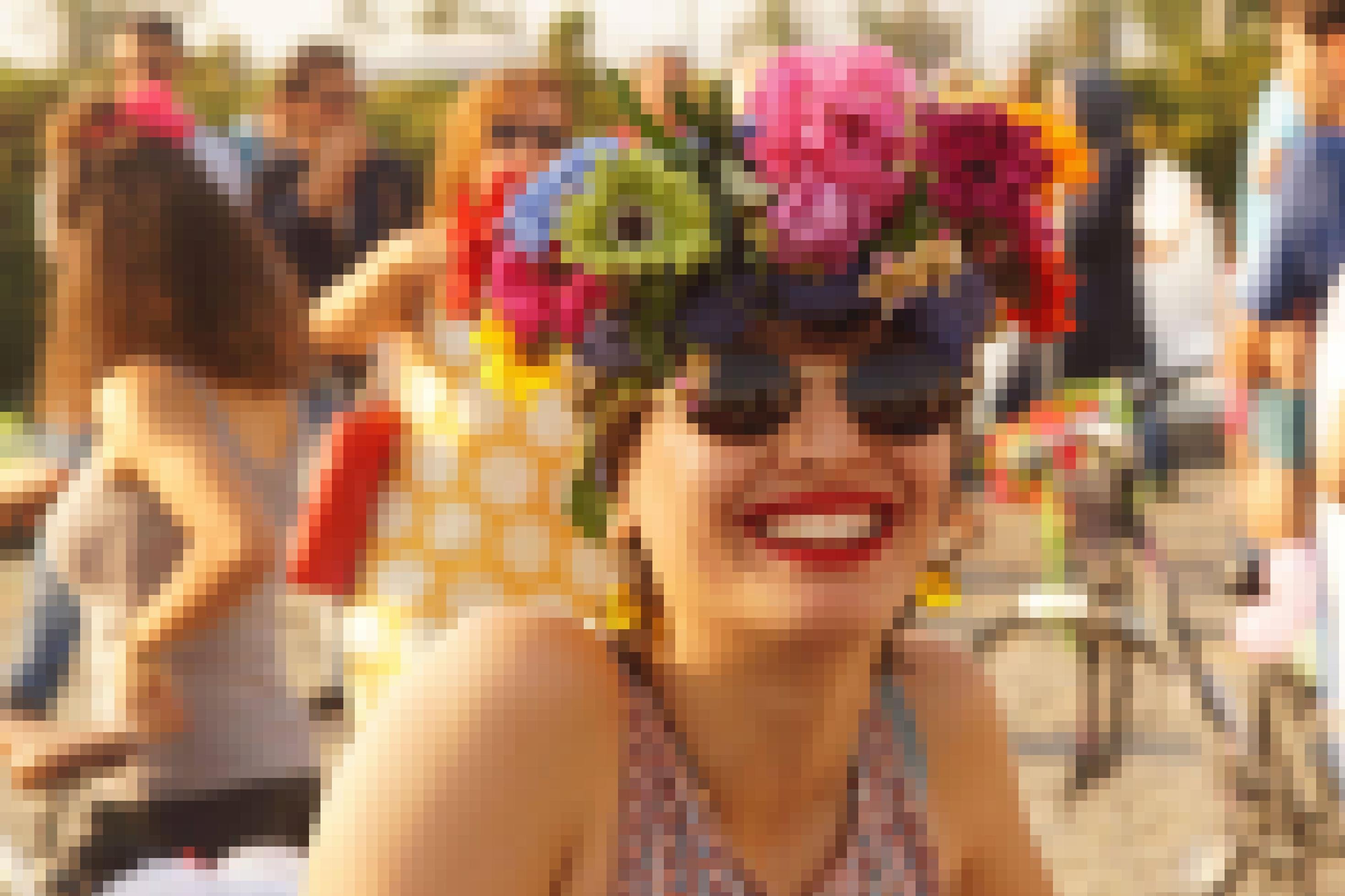 Die Portraitaufnahme zeigt eine Frau, die eine Sonnenbrille trägt und in die Kamera lacht. Sie hat sich ein Tuch um den Kopf gebunden und sich bunte Blumen ins Haar gesteckt. Im Hintergrund stehen weitere Teilnehmer der Ausfahrt.