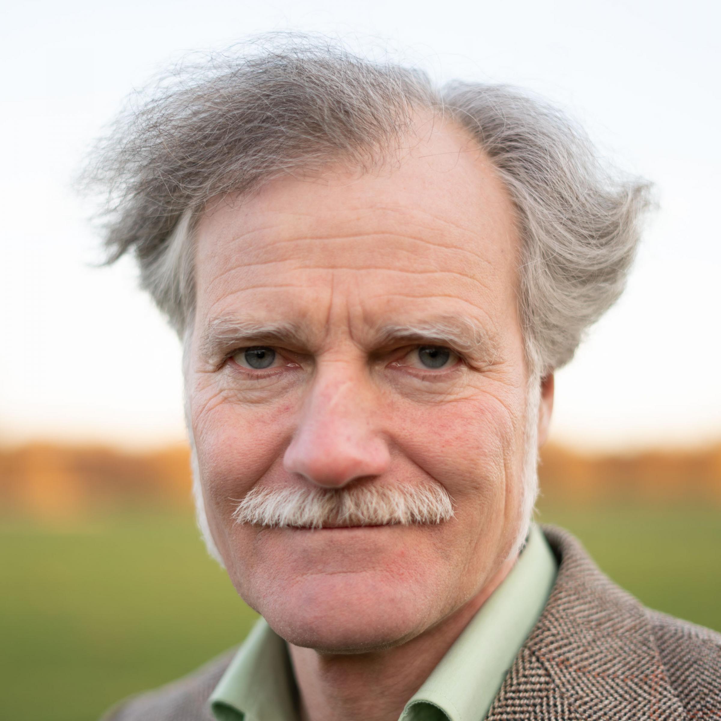 Der Field-Writer Gerhard Richter trägt eine grauen Schnurbart und die Haare als Seitenscheitel. Er trägt außerdem ein hellgrünes Hemd und darüber ein braunes Tweed-Sacko.