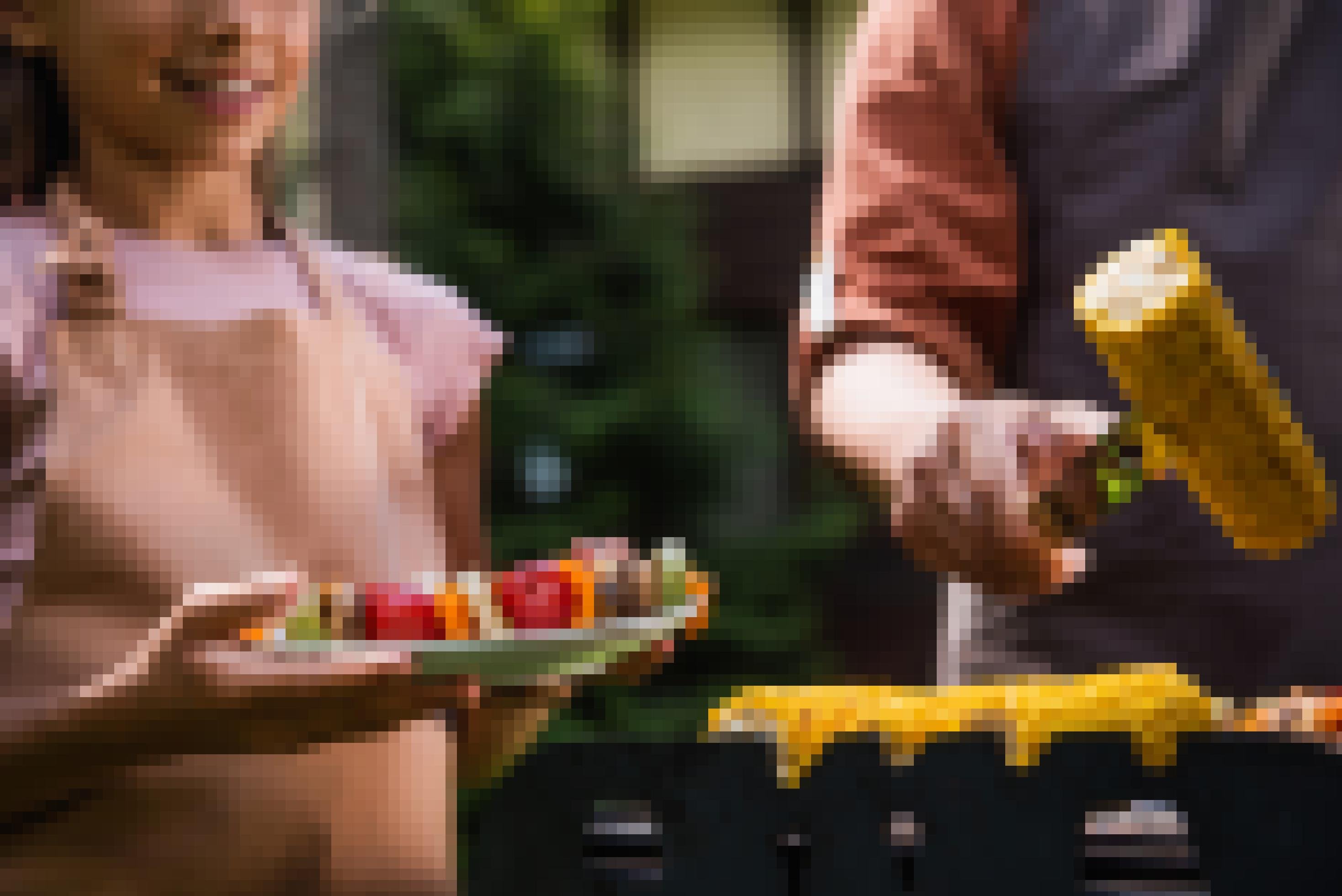Auf dem Grill im Vordergrund wendet ein Mann mit einer Grillzange den Maiskolben, neben ihm steht ein Mädchen mit einem Teller voller Gemüsespieße