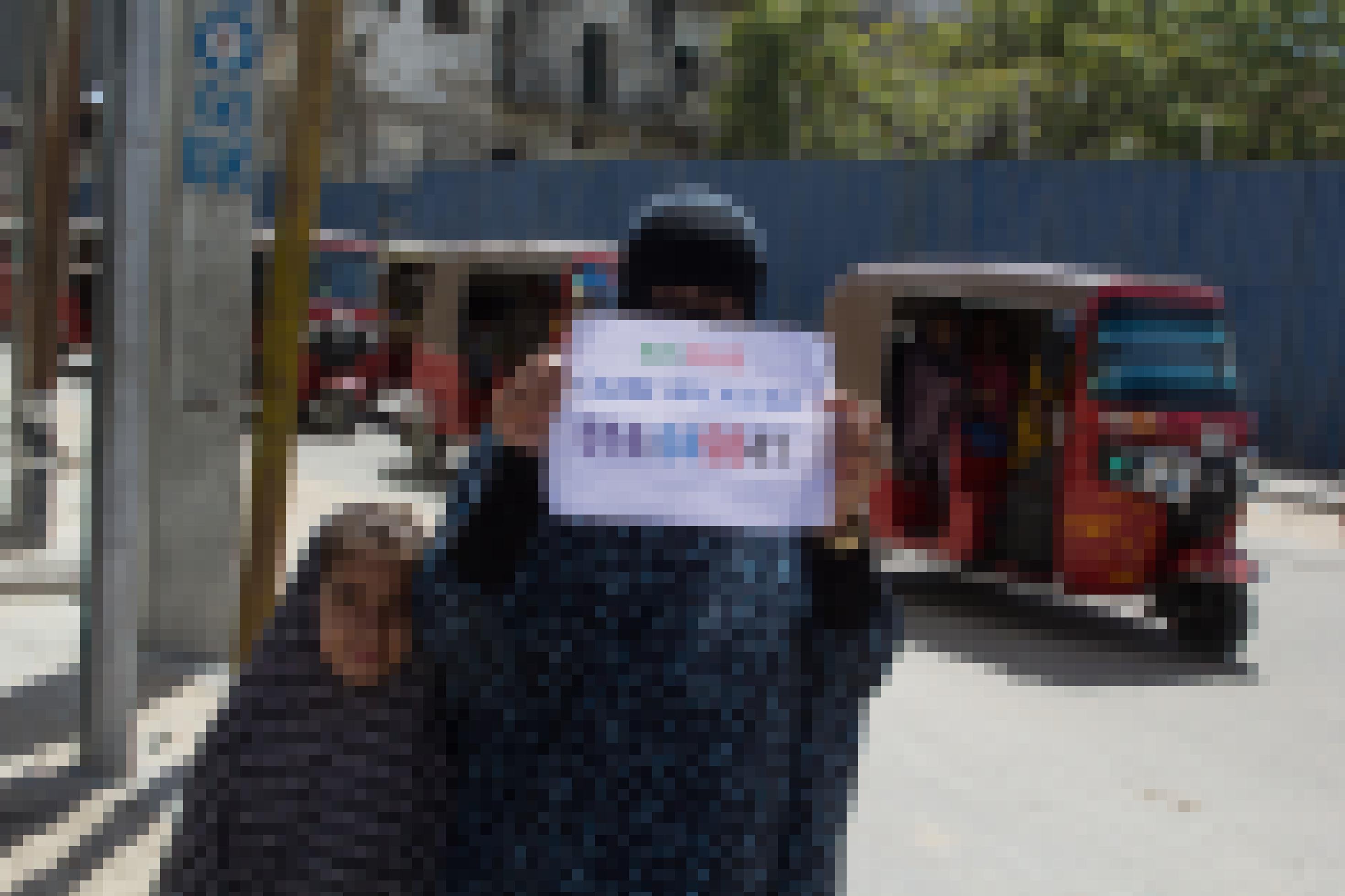 Eine verschleierte Frau und ihre Tochter stehen am Straßenrand, die Mutter hält einen Zettel mit Zahlen hoch. Im Hintergrund rote TukTuks.