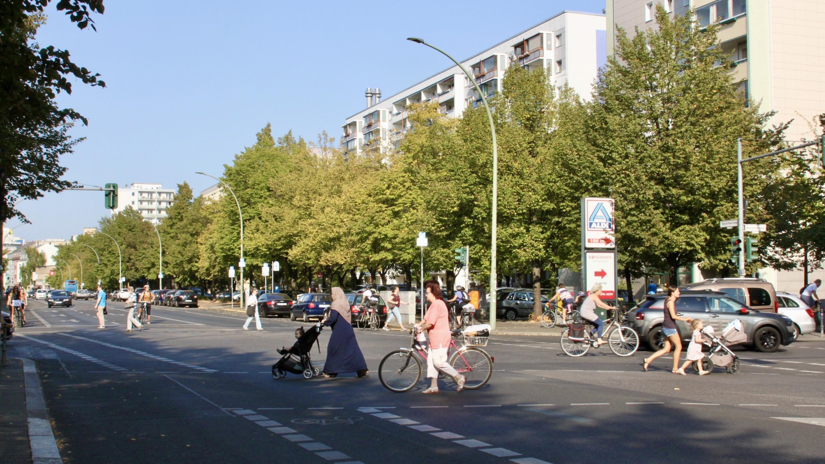 Eine breite Kreuzung in Berlin im Spätsommer. Die Sonne scheint. Zwei Frauen schieben Kinder in entgegengesetzter Richtung über die Ampel, eine weitere Frau schiebt ihr Fahrrad über die Kreuzung eine weitere fährt mit dem Rad im Hintergrund über die Kreuzung.