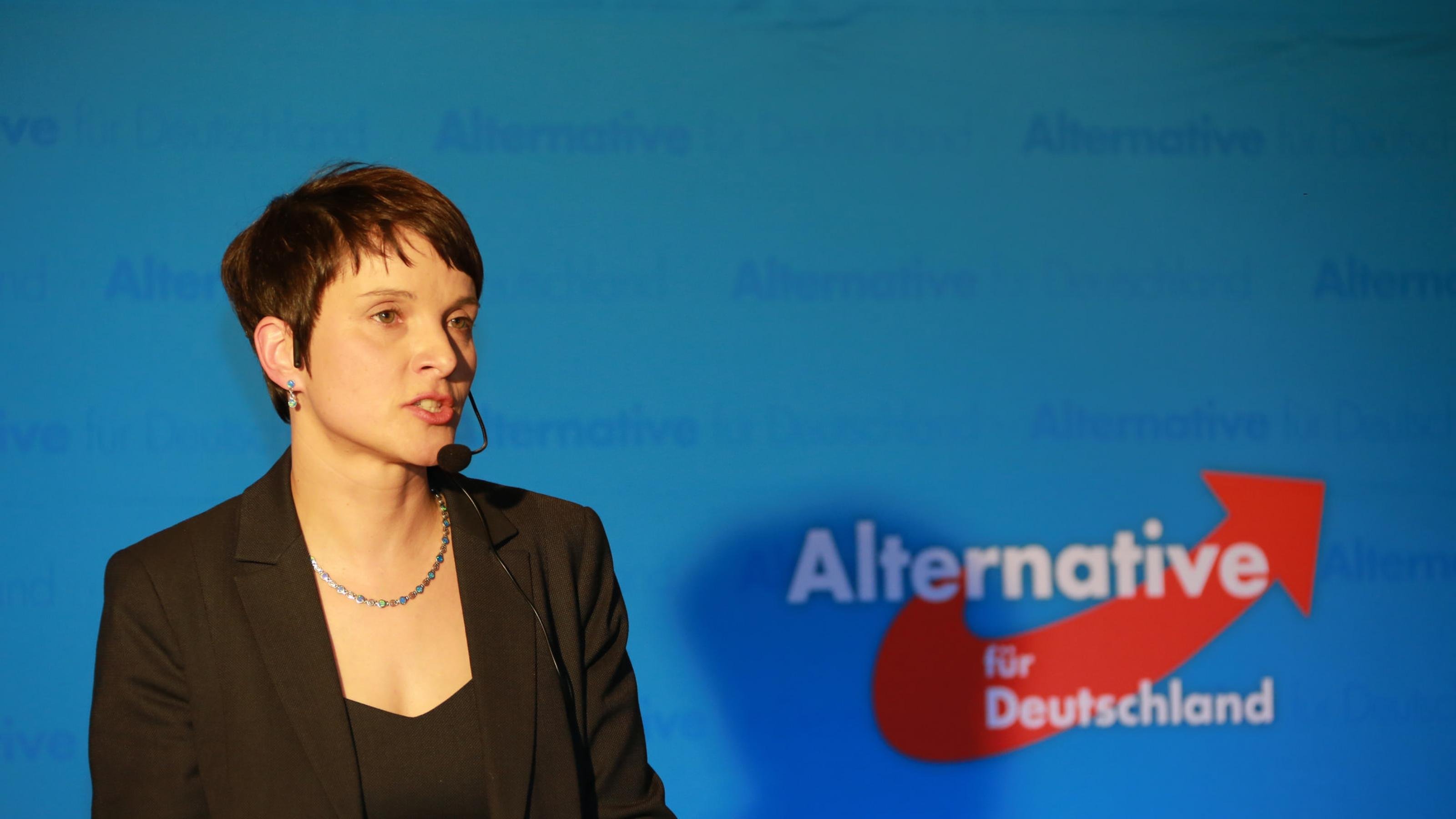 Frauke Petry vor einer blauen Wand mit AfD-Logo.
