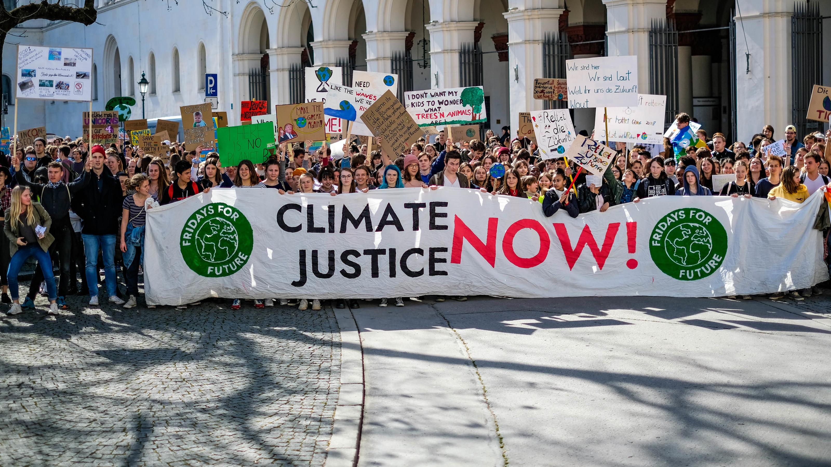 Junge Leute aus der FridaysForFuture-Bewegung stehen vor einem Gebäude der Universität München und halten ein Banner mit der Aufschrift „Climate Action Now“ vor sich.