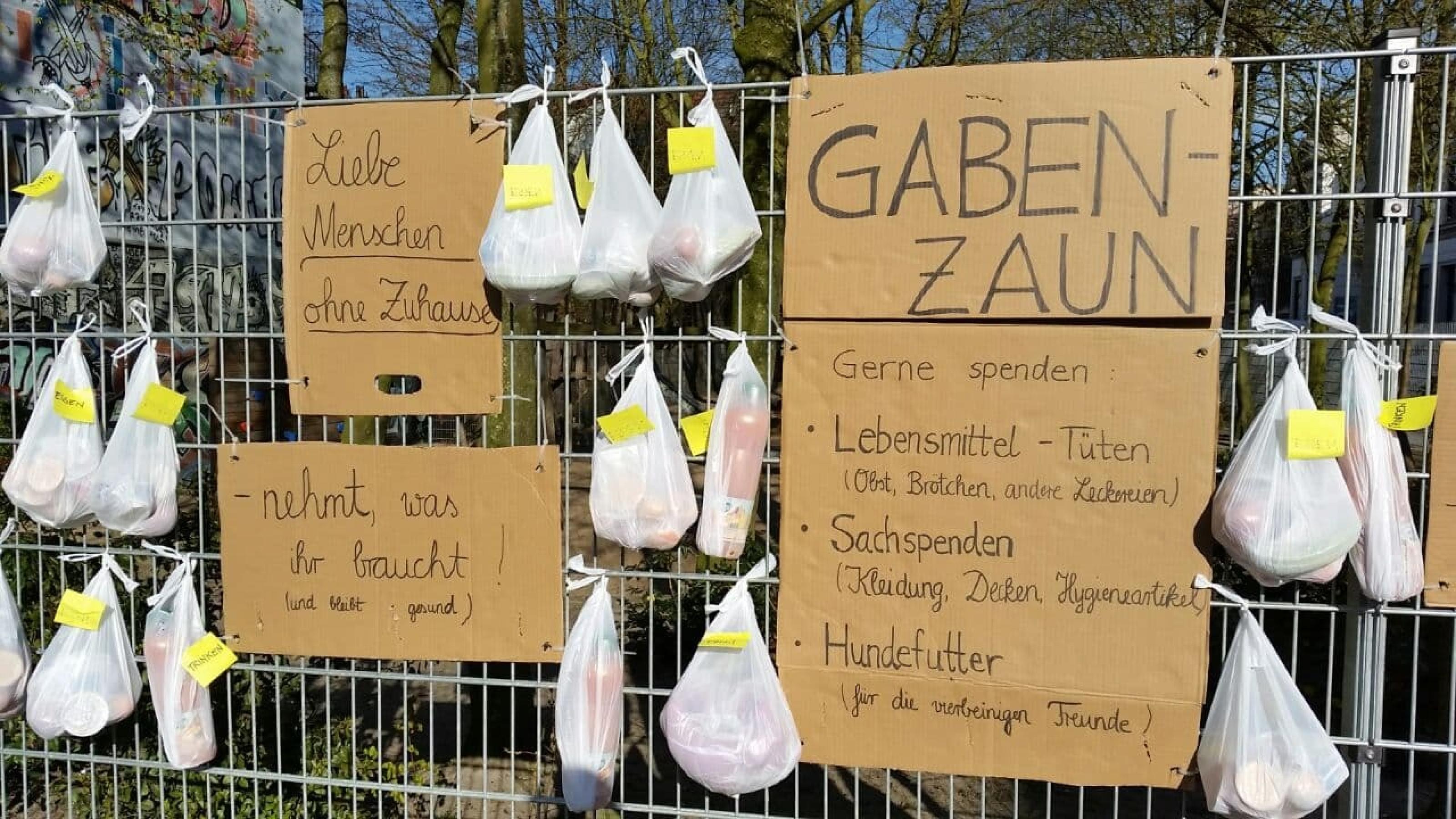 An einem hohen Gitterzaun hängen weiße Plastiktuten mit Lebensmitteln und Pappschilder, die zum Spenden und Mitnehmen aufrufen.