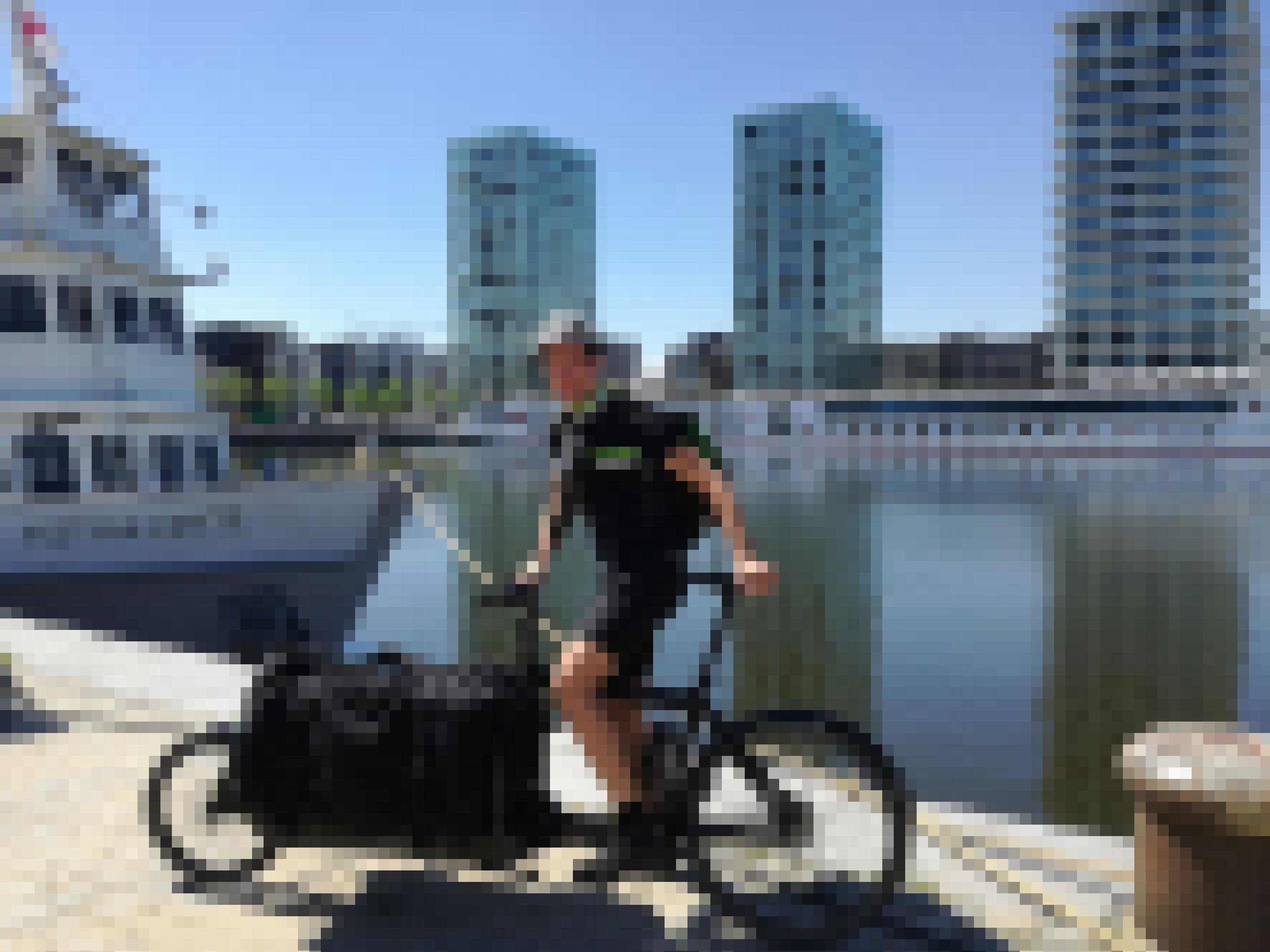 Ein sportlicher Radfahrer in schwarzer Kleidung auf steht mit seinem Lastenrad vor einem Gewässer in der Stadt.