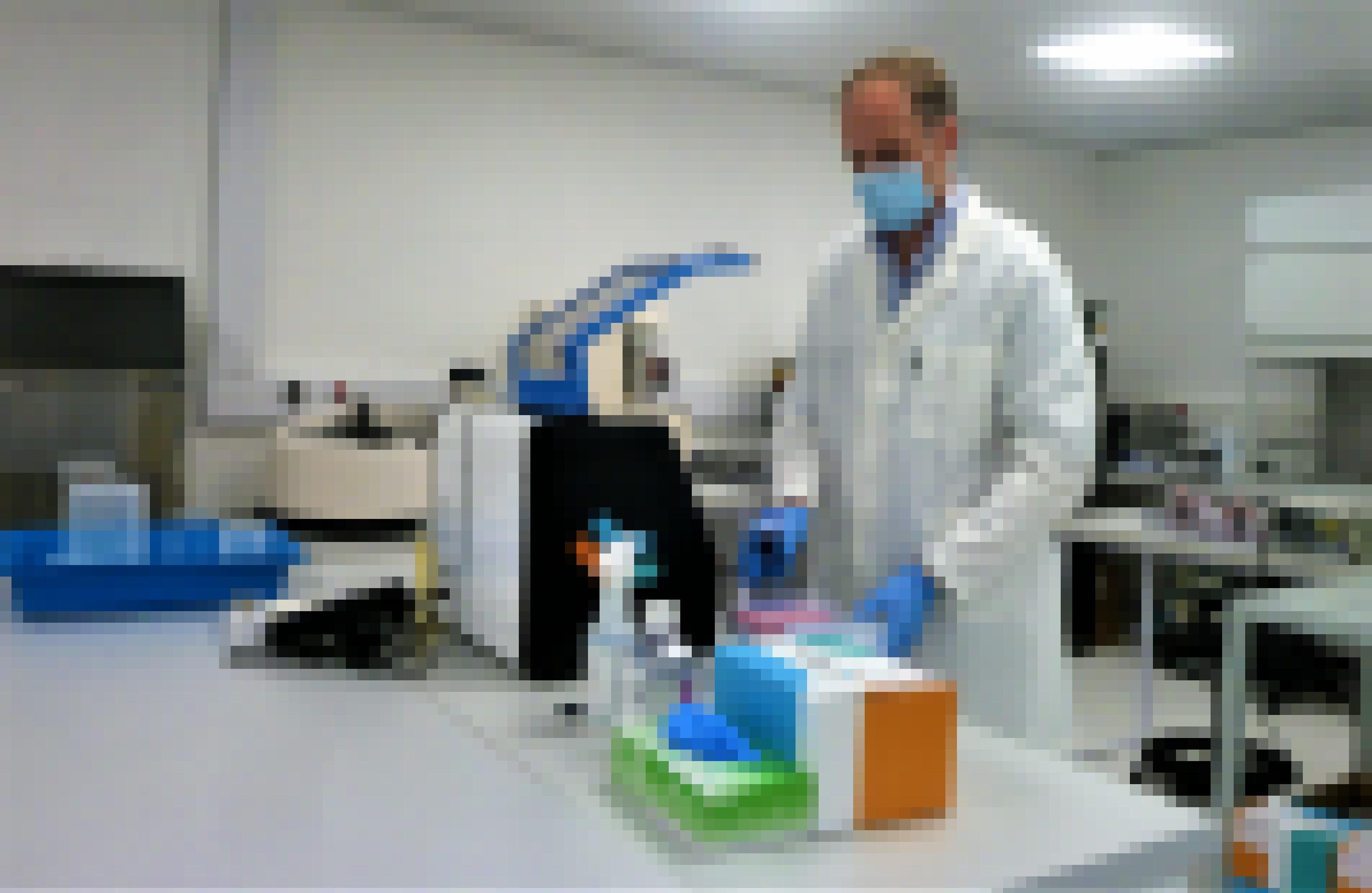 Der Wissenschaftler steht in weißem Kittel und mit Gesichtsmaske im Labor. Neben einem Gerät vor ihm stehen eine Sprühflasche und ein Karton mit Einmalhandschuhen.
