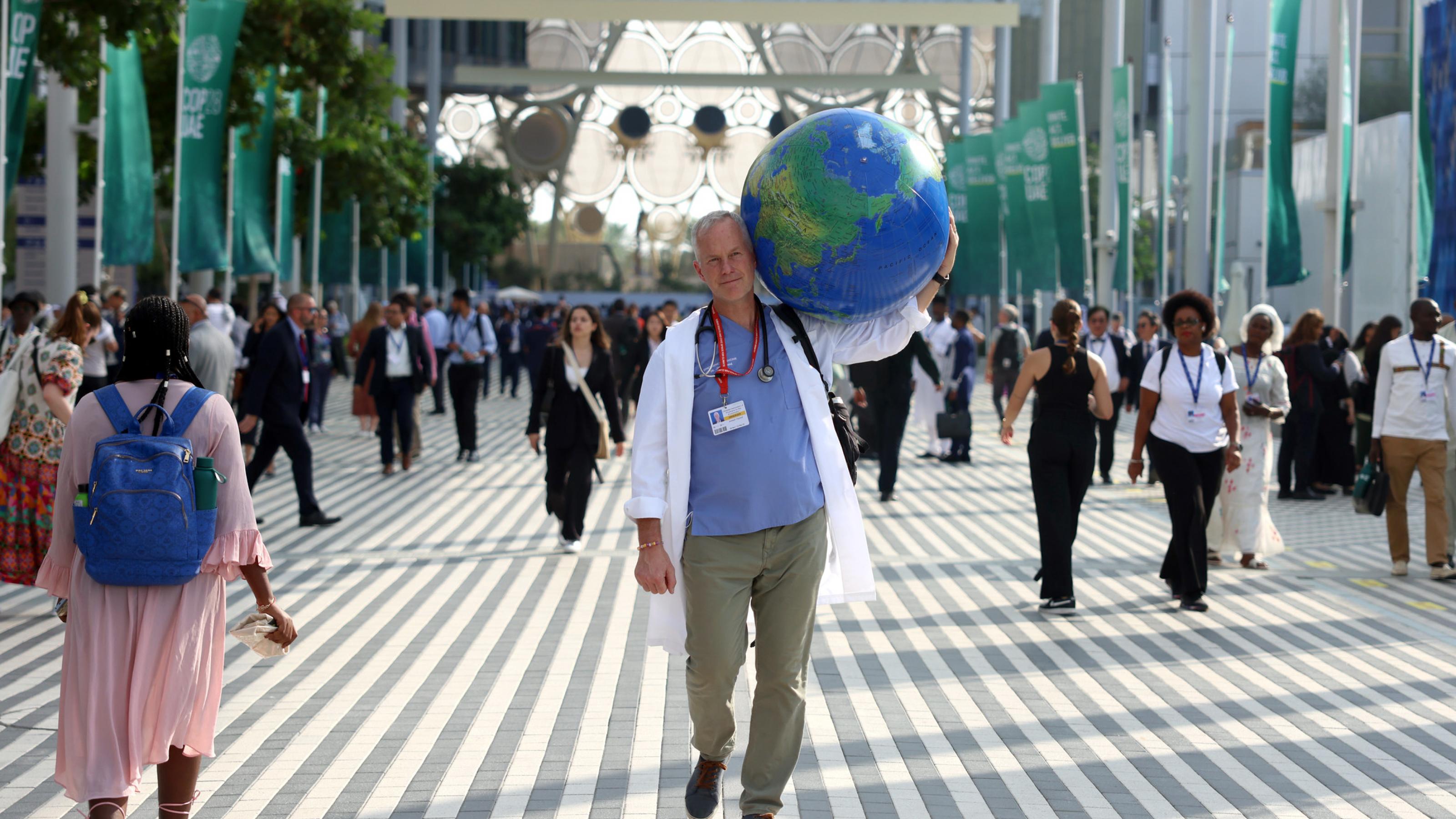 Arzt in Medizinkittel mit Stethoskop um den Hals trägt symbolisch die Erde (als aufgeblasener Wasserball) auf seiner Schulter, während er über das gut besuchte COP28-Gelände in Dubai geht