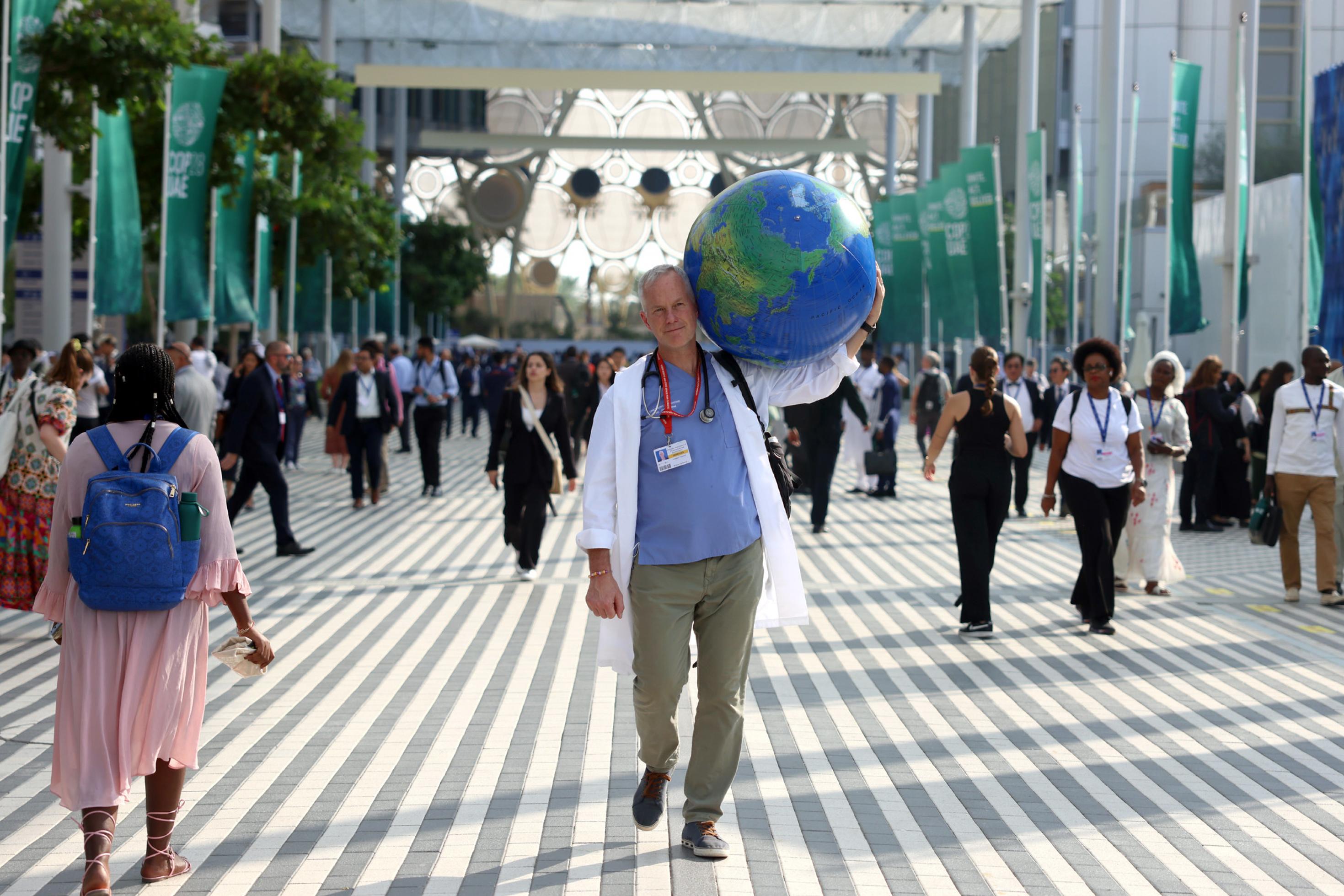 Arzt in Medizinkittel mit Stethoskop um den Hals trägt symbolisch die Erde (als aufgeblasener Wasserball) auf seiner Schulter, während er über das gut besuchte COP28-Gelände in Dubai geht