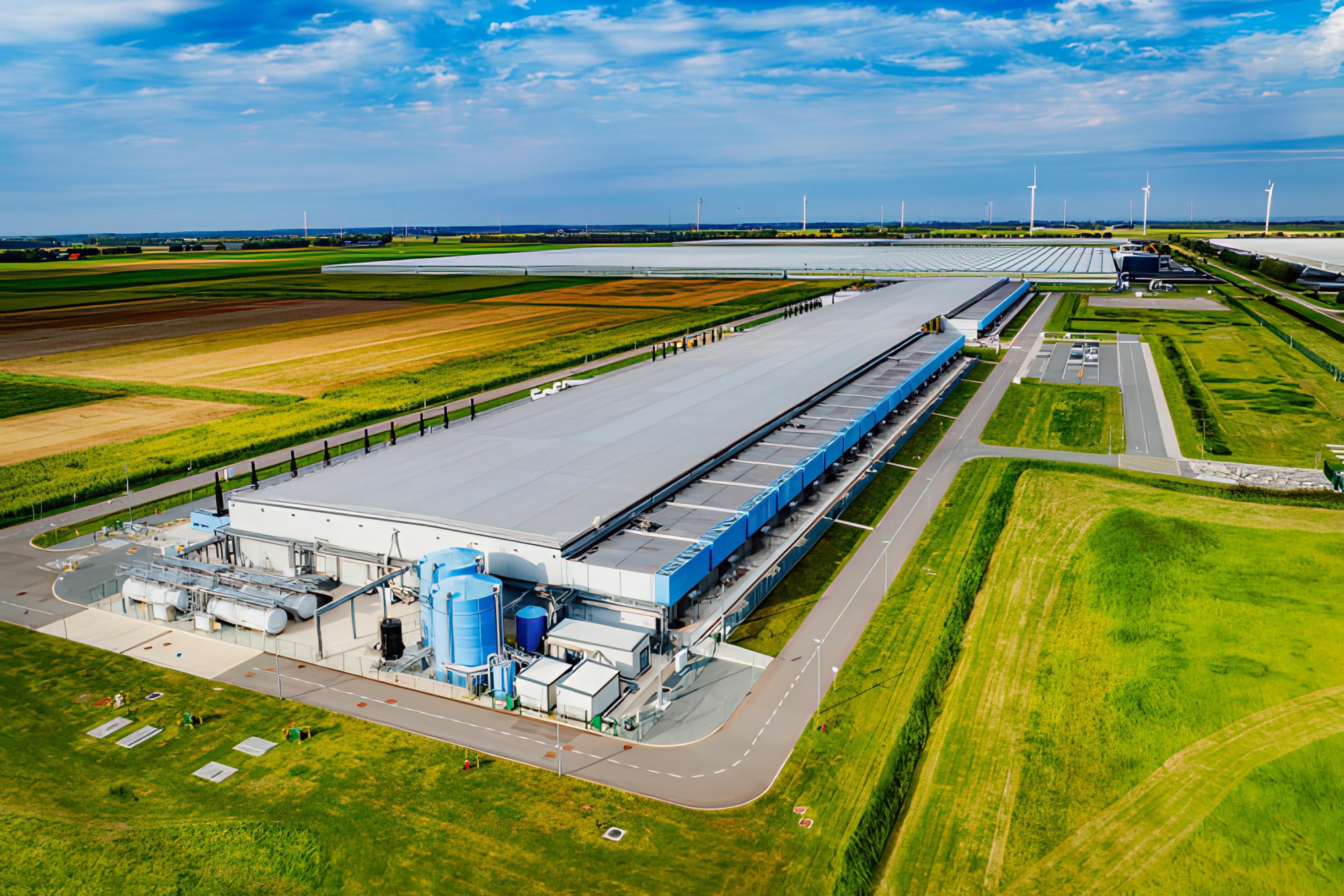 Moderne Rechenzentren sehen aus wie Fabriken. Das Bild zeigt ein langgestrecktes, flaches Gebäude mit technischer Infrastruktur in einer unbebauten Umgebung. Es ist ein Rechenzentrum des Internetgiganten Google im niederländischen Middenmeer.