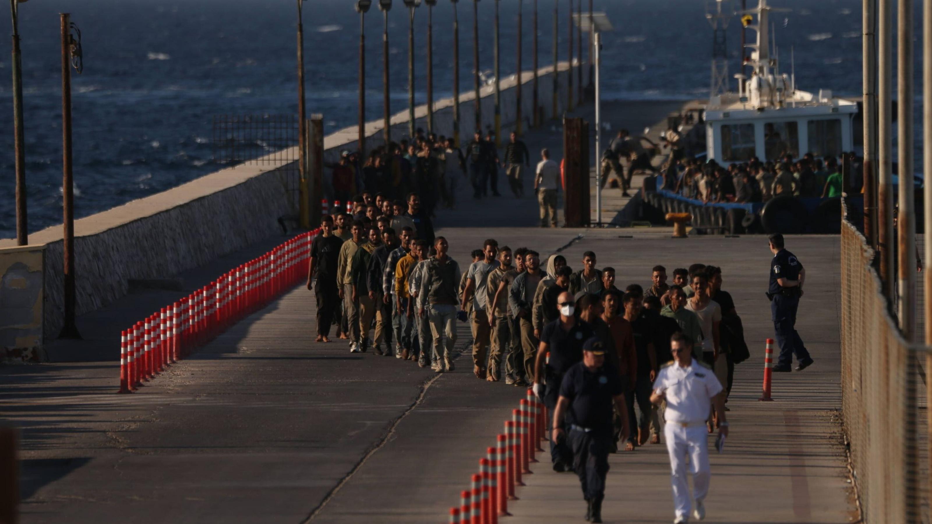 Eine lange Schlange von Menschen läuft barfuß oder in einfacher Kleidung hintereinander auf einem Pier am Meer. Sie werden von Polizisten begleitet. Im Hintergrund liegt ein Schiff, auf dem sich weitere Menschen befinden. Die Szene wirkt angespannt und zeigt Migranten, die in Griechenland angekommen sind.