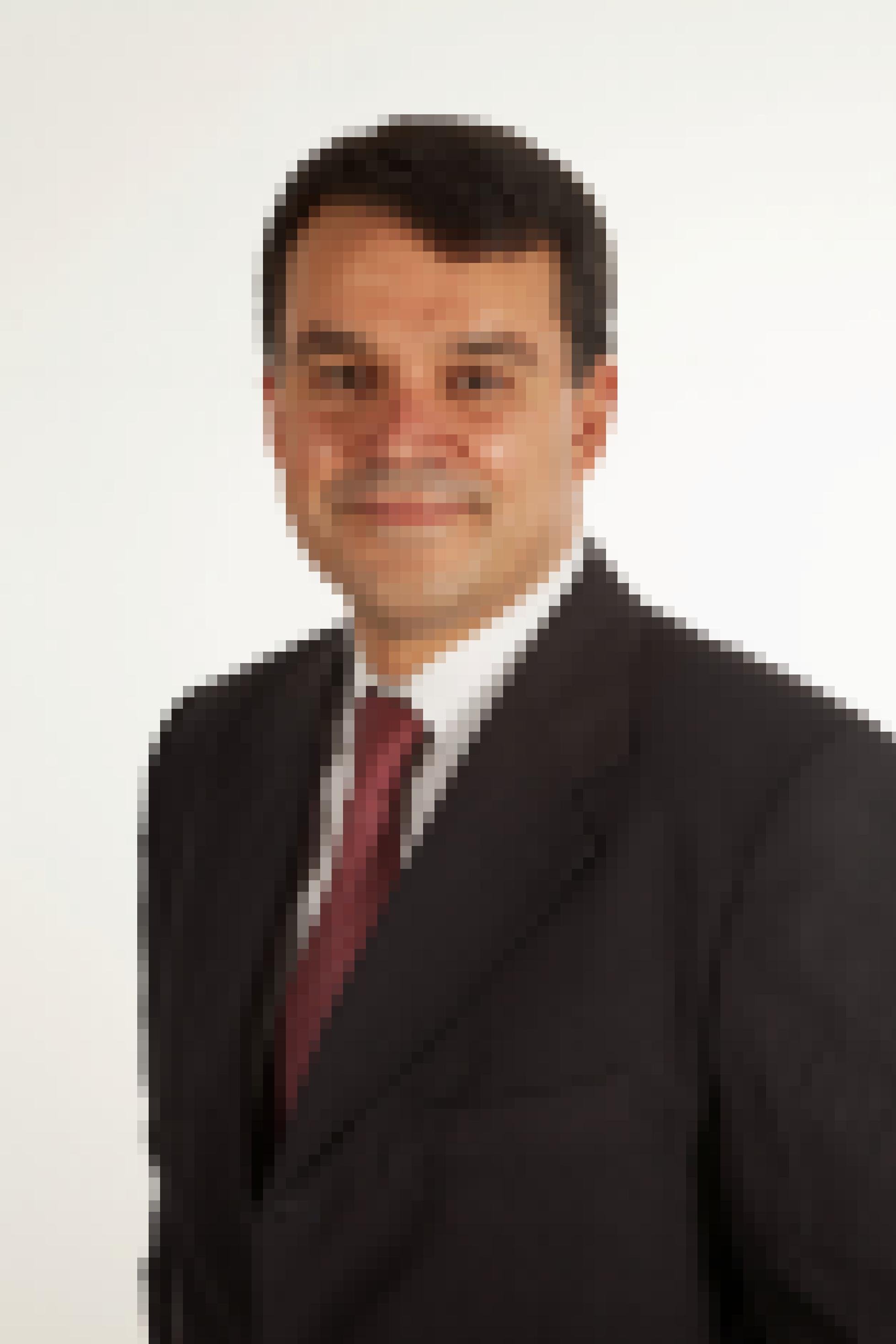 Ein Portraitbild eines Mannes, der in leicht seitlicher Haltung zu sehen ist. Er grinst und trägt ein schwarzes Sakko. Darunter ist ein weißes Hemd und eine rote Krawatte zu sehen.