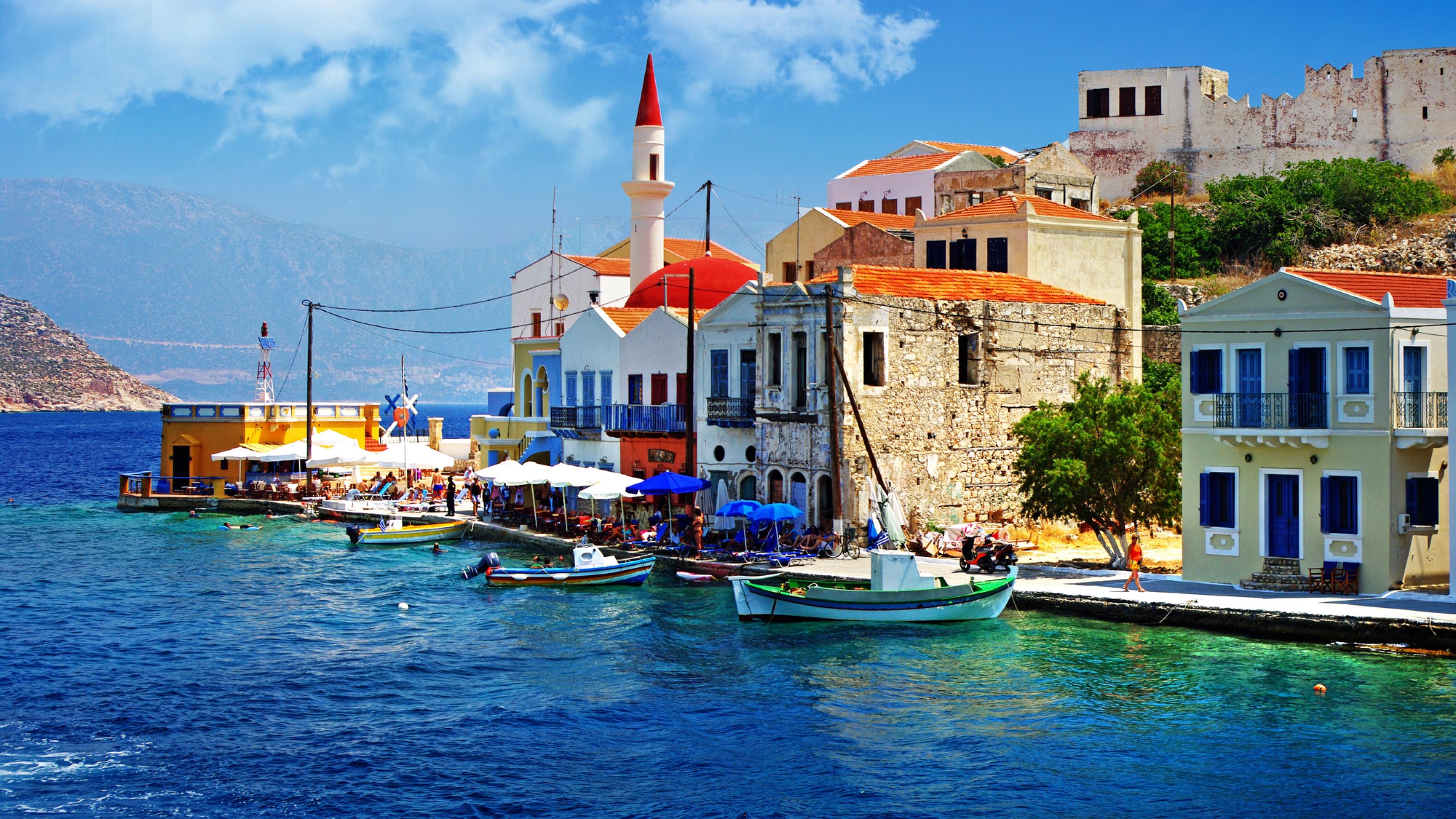 Die Szene zeigt eine lebendige Hafenpromenade auf einer griechischen Insel. Entlang des Wassers stehen Häuser mit roten Ziegeldächern, weißen und gelben Fassaden sowie blauen Türen und Fensterläden. Vor den Häusern befinden sich Cafés und Tavernen mit Sonnenschirmen, unter denen Menschen entspannen. Bunte Fischerboote schaukeln im klaren, türkisblauen Wasser. Im Hintergrund rahmen kahle Hügel und ein wolkenloser Himmel die malerische Szene ein.