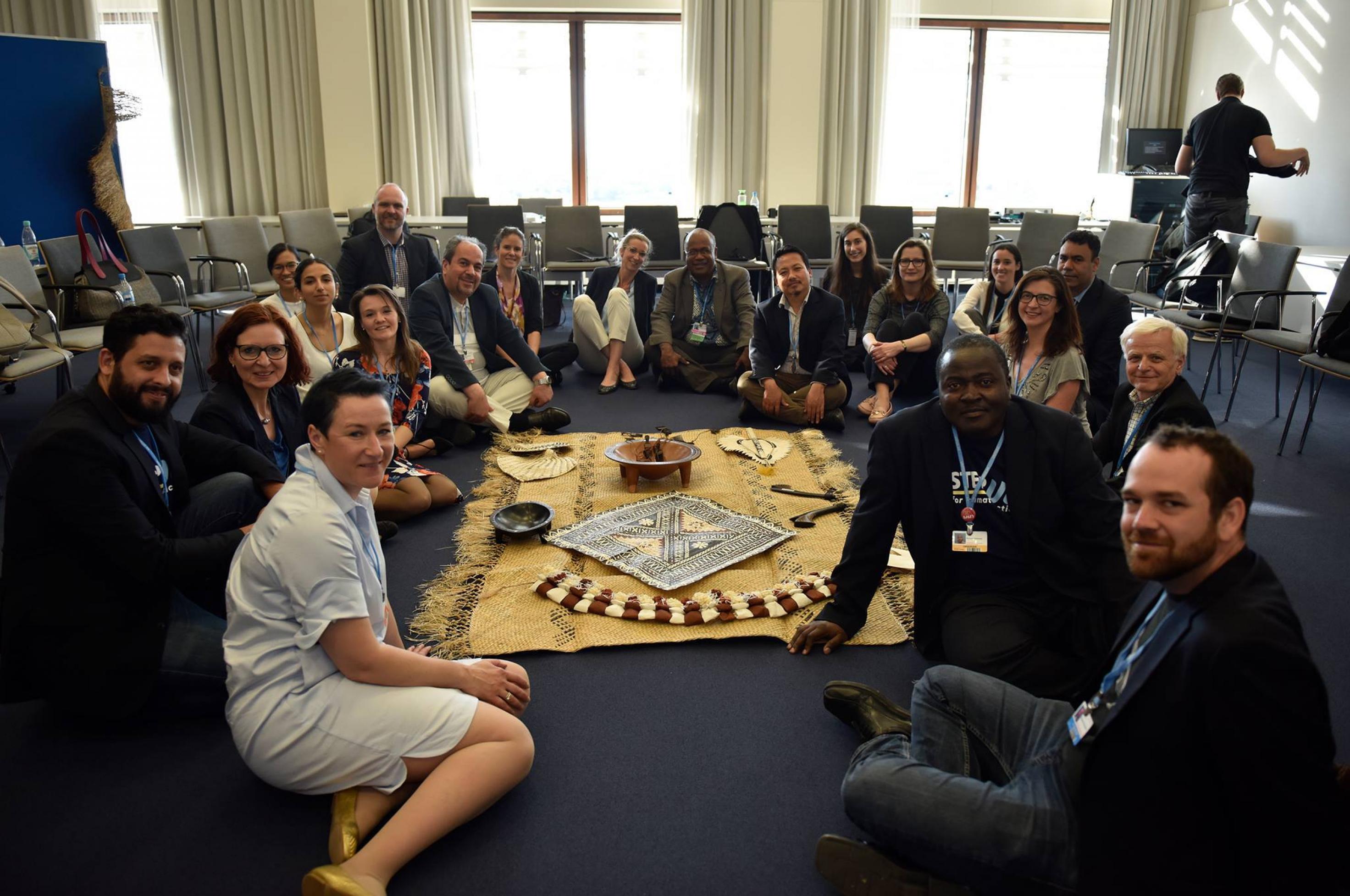 Eine internationale Gruppe von Frauen und Männern sitzt in einem Konferenzraum auf dem Boden und lächelt in die Kamera. Sie bereiten sich darauf vor, einen Dialog nach dem Talanoa-Format zu führen. Sie sitzen rund um eine geflochtene Matte mit einem Gefäß für Kava-Sud. Allerdings nur für das Foto – beim eigentlichen Dialog saßen die Teilnehmer auf den Stühlen im Hintergrund.