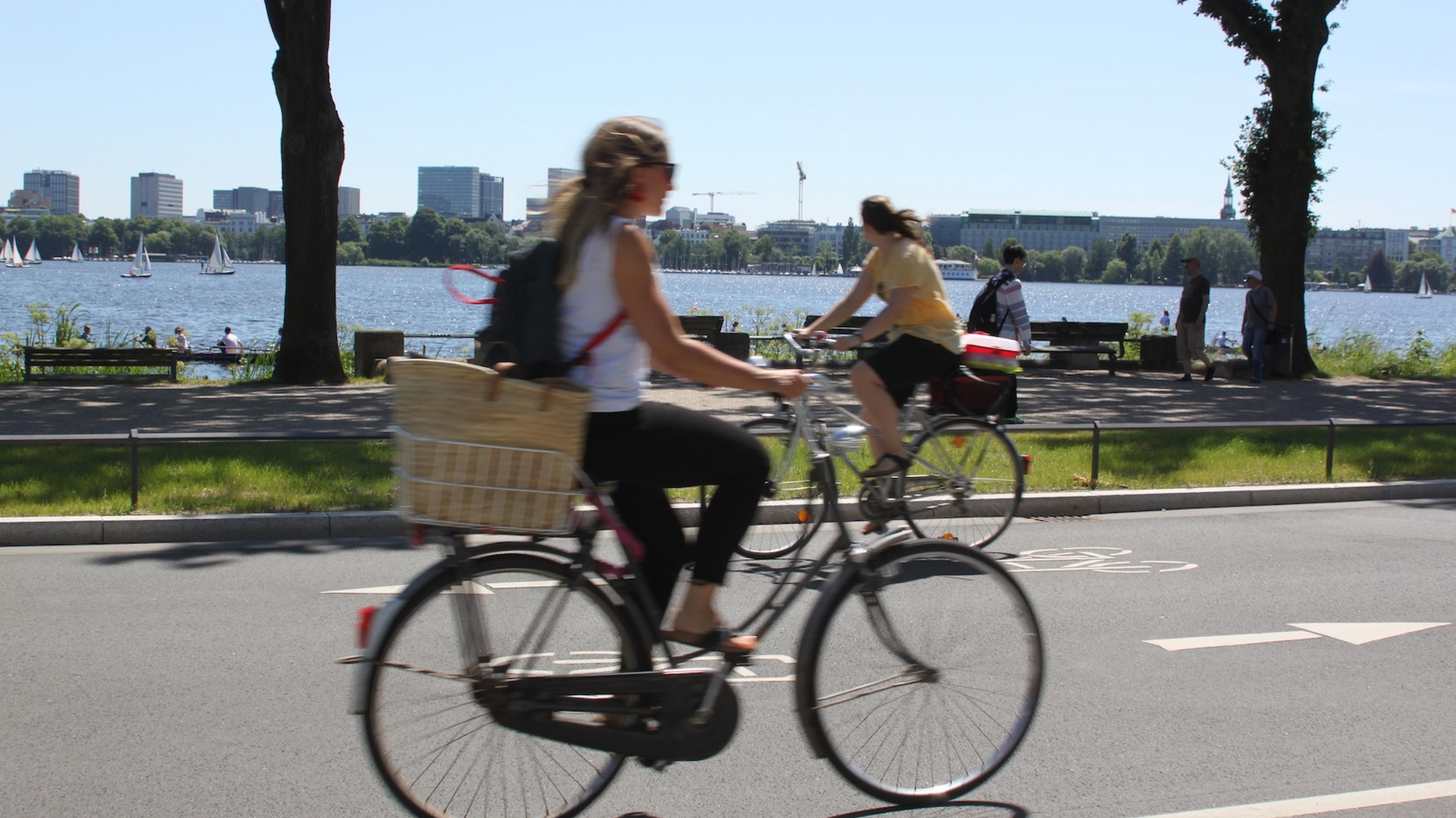 Zwei Radfahrerinnen fahren in jeweils entgegengesetzter Richtung aneinander vorbei. Das Wetter ist sonnig.