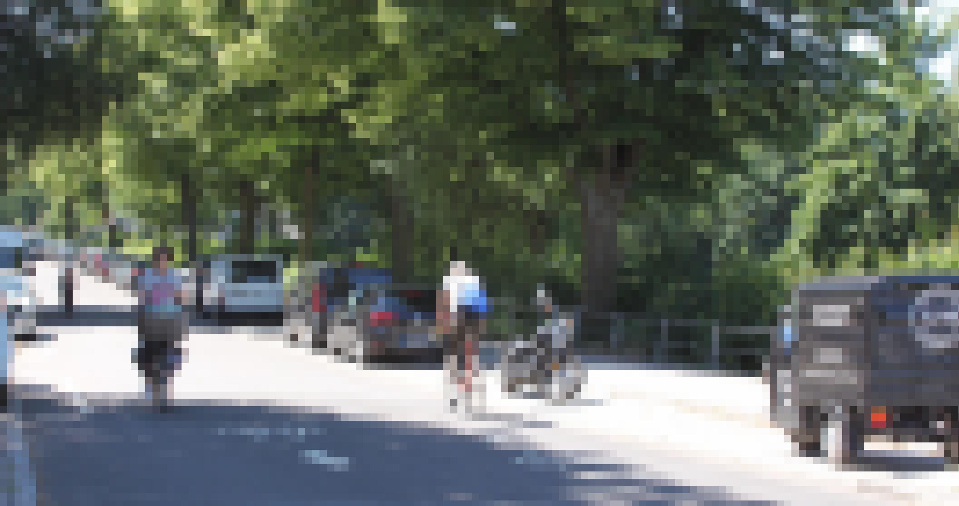 Drei Radfahrer fahren bei sonnigem Wetter auf einer Straße, die von grünen Bäumen gesäumt ist. An beiden Seiten parken Autos und ein Motorrad.