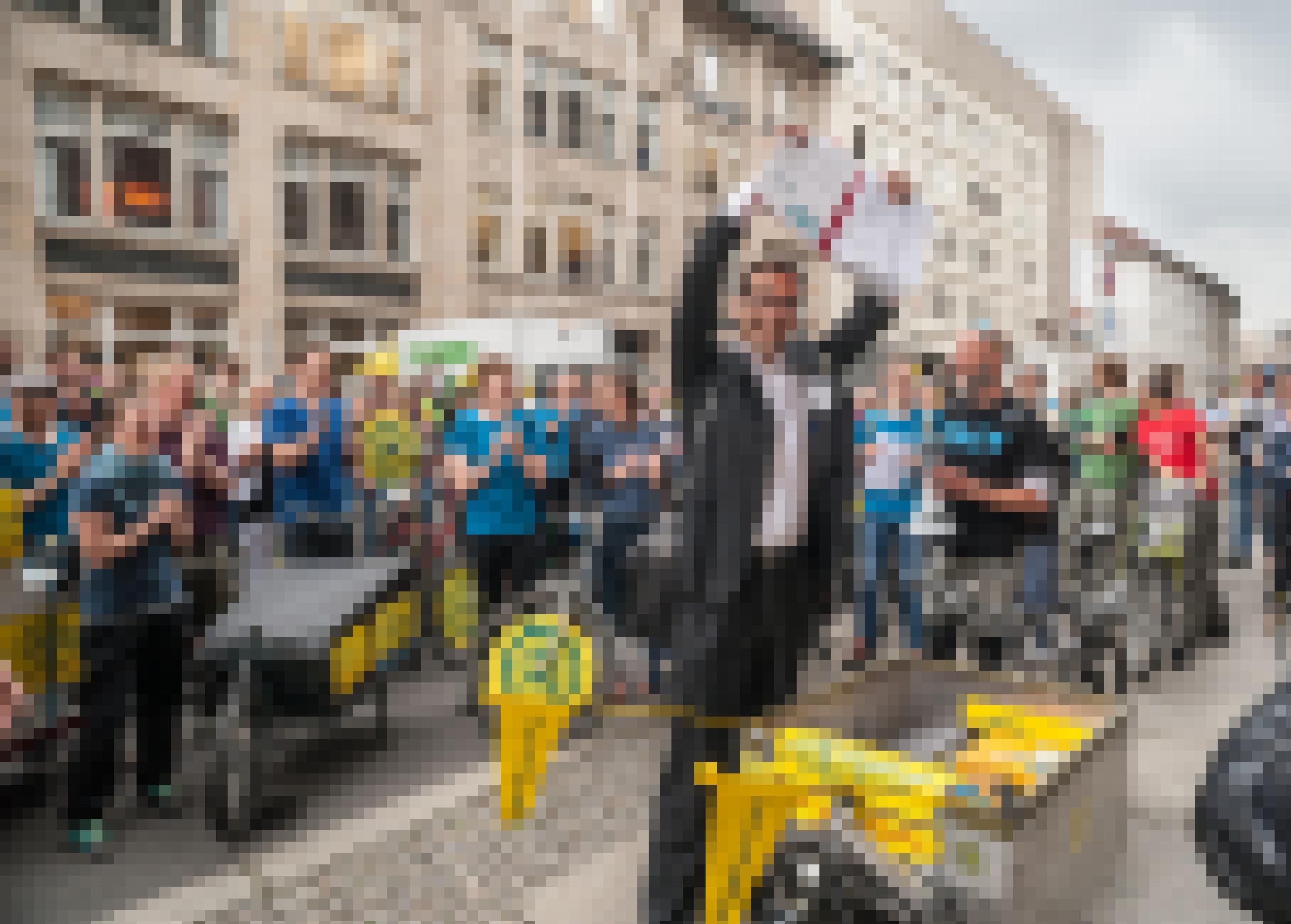 Eine große Menschenmenge in vorwiegend hellblauen T-Shirts steht auf der Straße vor einer Häuserfront. Im Vordergrund steht ein Mann in Anzug, der ein geöffnetes Buch hoch über seinen Kopf hält und in die Kamera lächelt.