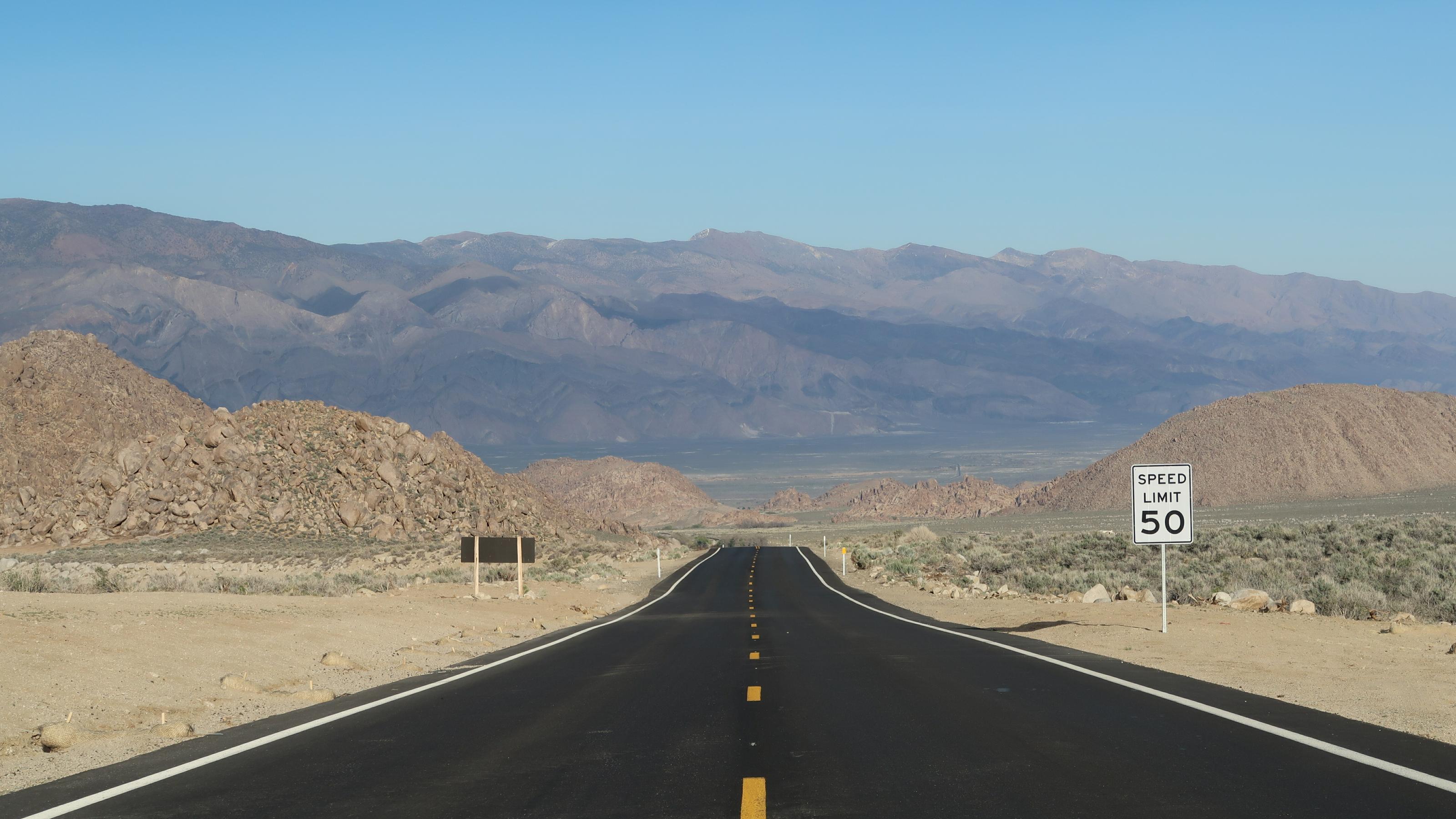 Das Bild zeigt eine leere Straße, die in die Ferne führt. Neben der Straße liegt eine Wüste, in der Ferne sind Berge zu sehen.