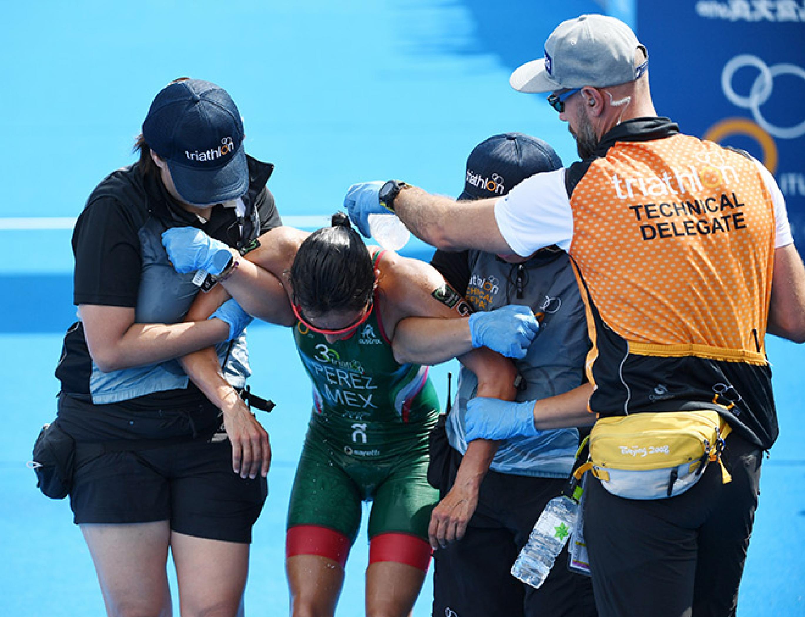 Eine Läuferin mit gesenktem Kopf wird von zwei Frauen gestützt, während ein Mann mit offizieller Weste ihr zur Kühlung Wasser über Schultern und Nacken gießt. Nach dem Frauen-Triathlon im August 2019 brauchte die Sportlerin Hilfe.