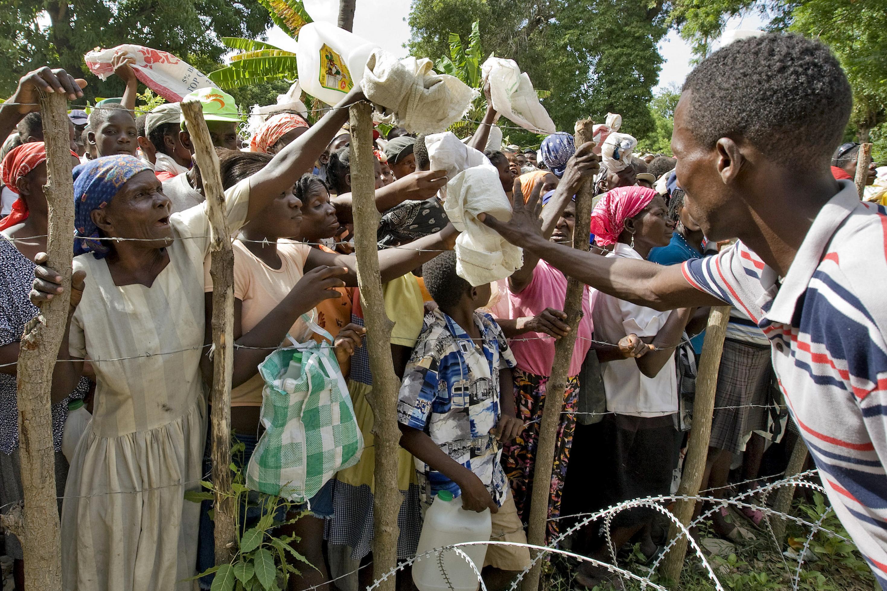 Ein Mitarbeiter einer UN-Hilfsorganisation sammelt Beutel von einer Gruppe von Frauen und Kindern hinter einem Stacheldraht-Zaun ein, die 2008 bei Port-au-Prince auf Haiti auf Nahrungsmittelhilfe warten. Das Fotoarchiv der Vereinten Nationen hat eine große Anzahl solcher Bilder von menschlichem Leid von allen Kontinenten.