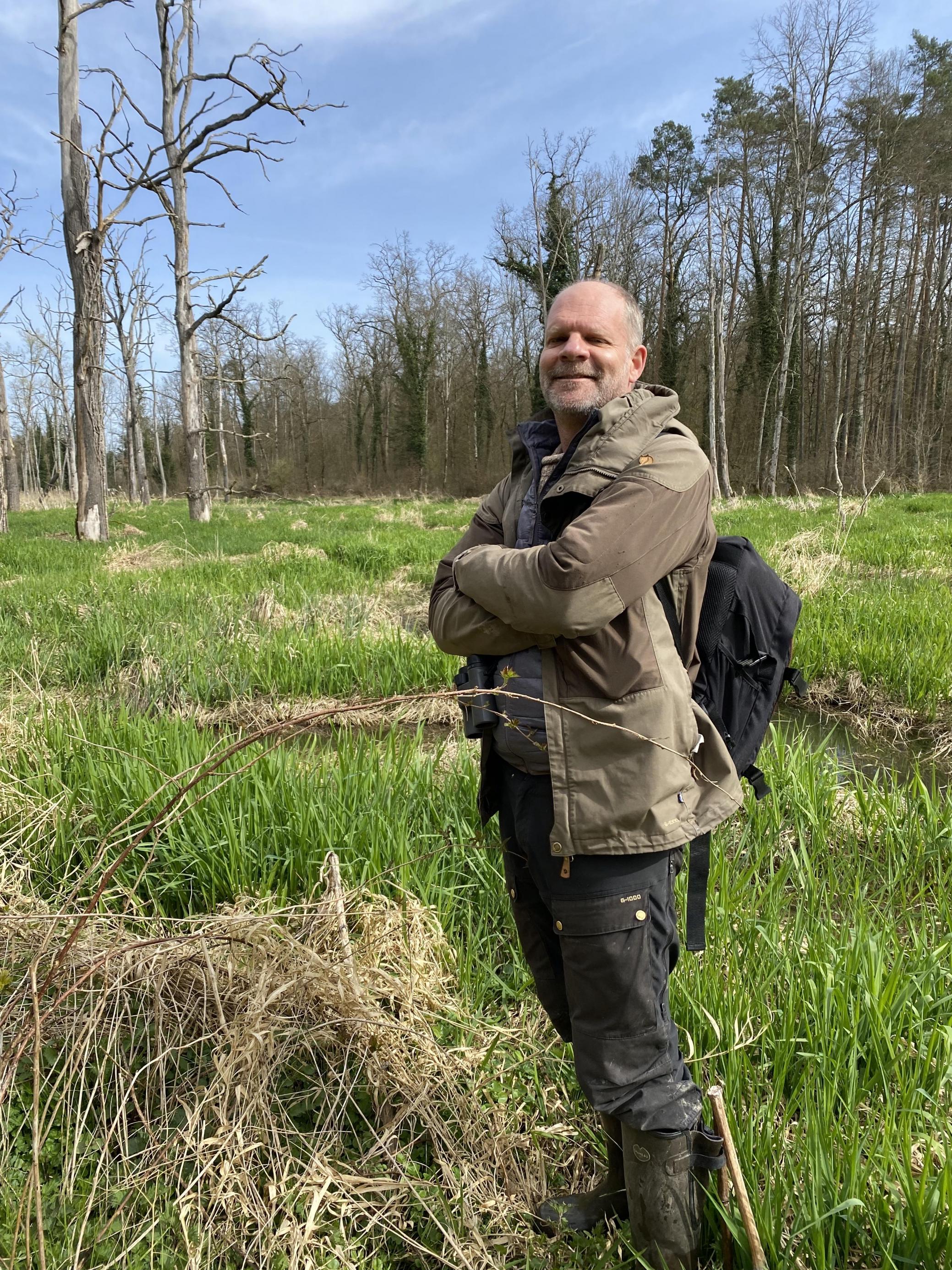 Christof Angst steht mit verschränkten Armen und lachend auf einer sumpfigen Wiese. Er trägt einen Rucksack und eine Outdoor-Jacke.