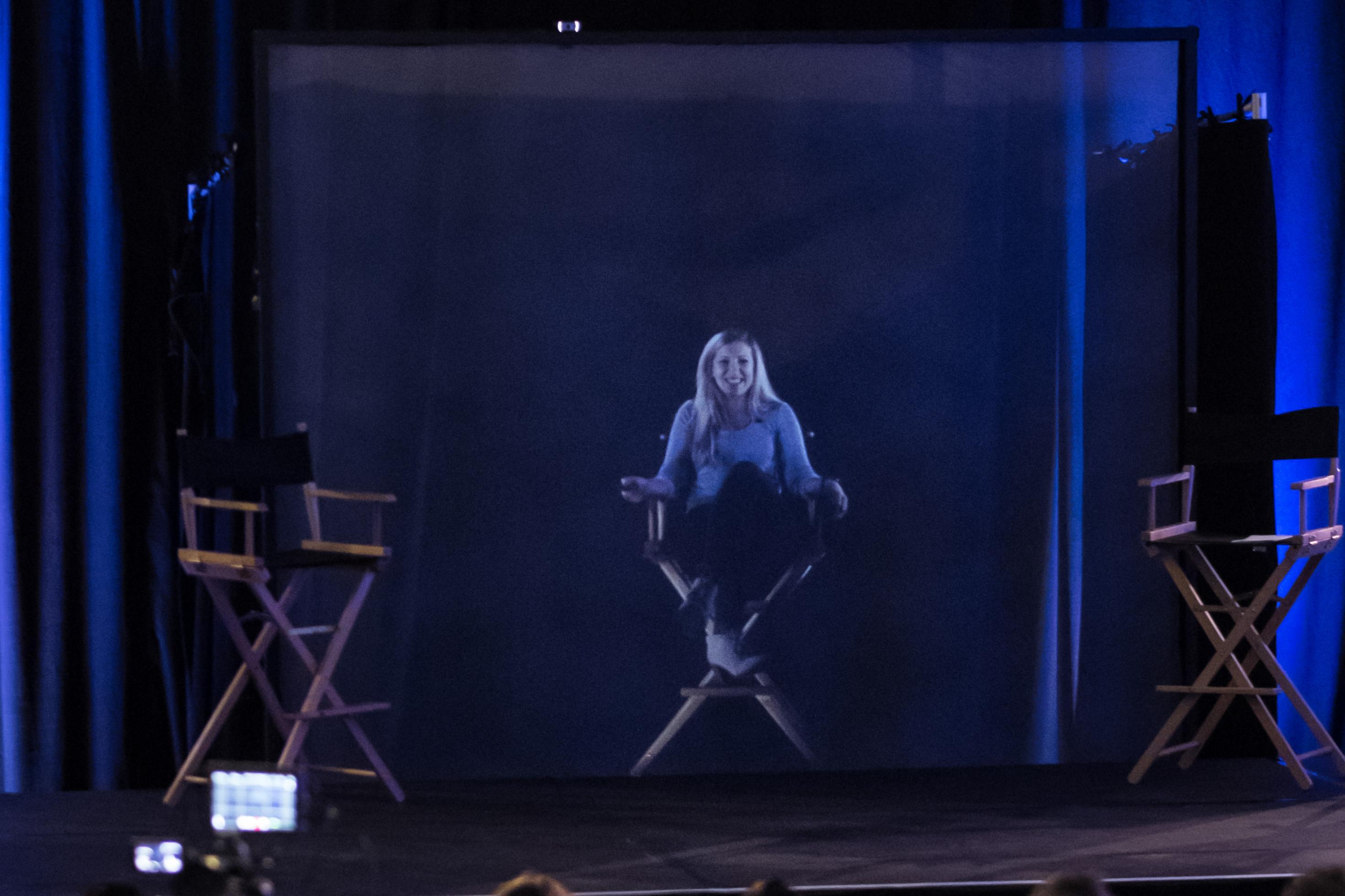 Auf einem Podium sitzt eine Frau auf einem hohen Stuhl. Scheinbar. Denn tatsächlich ist sie nicht in Person anwesend, sondern wurden nur als Hologramm zugeschaltet.