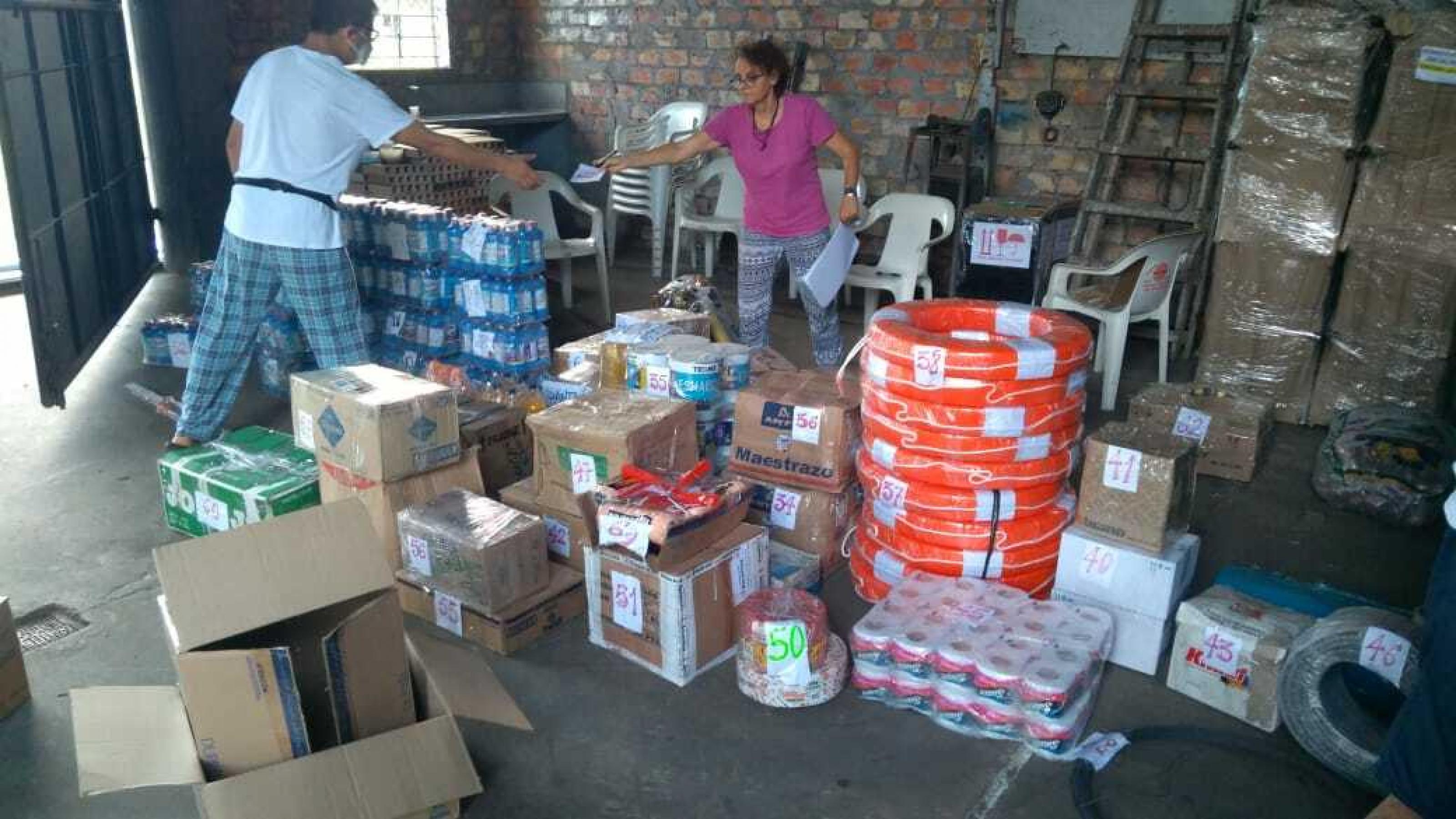 Ein Lager mit Wasserflaschen, Klopapier und Kartons, ein Mann und eine Frau stellen die Waren zusammen.