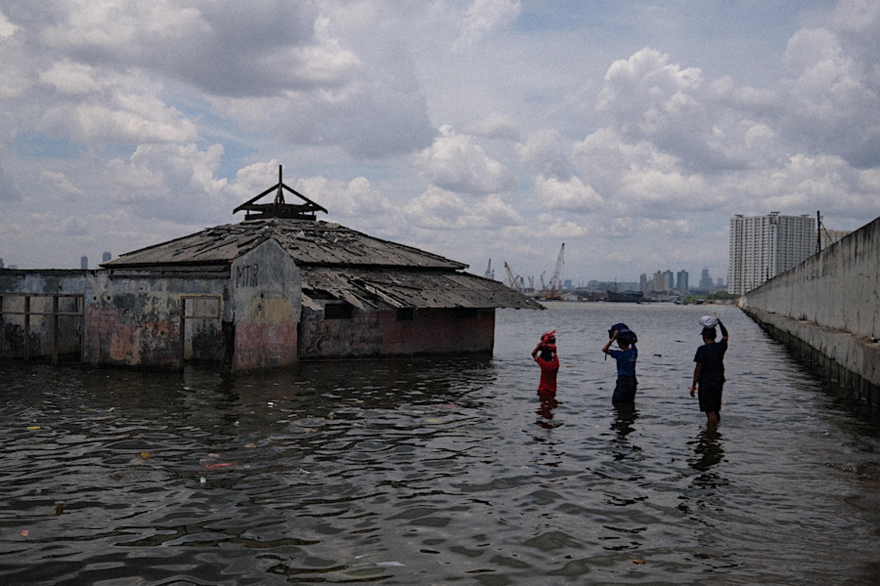 Drei Personen waten zu einer Moschee, die halb versunken  im Meer liegt. Dahinter ist die riesige Schutzmauer zu sehen, die Jakarta vor den steigenden Fluten schützen soll.