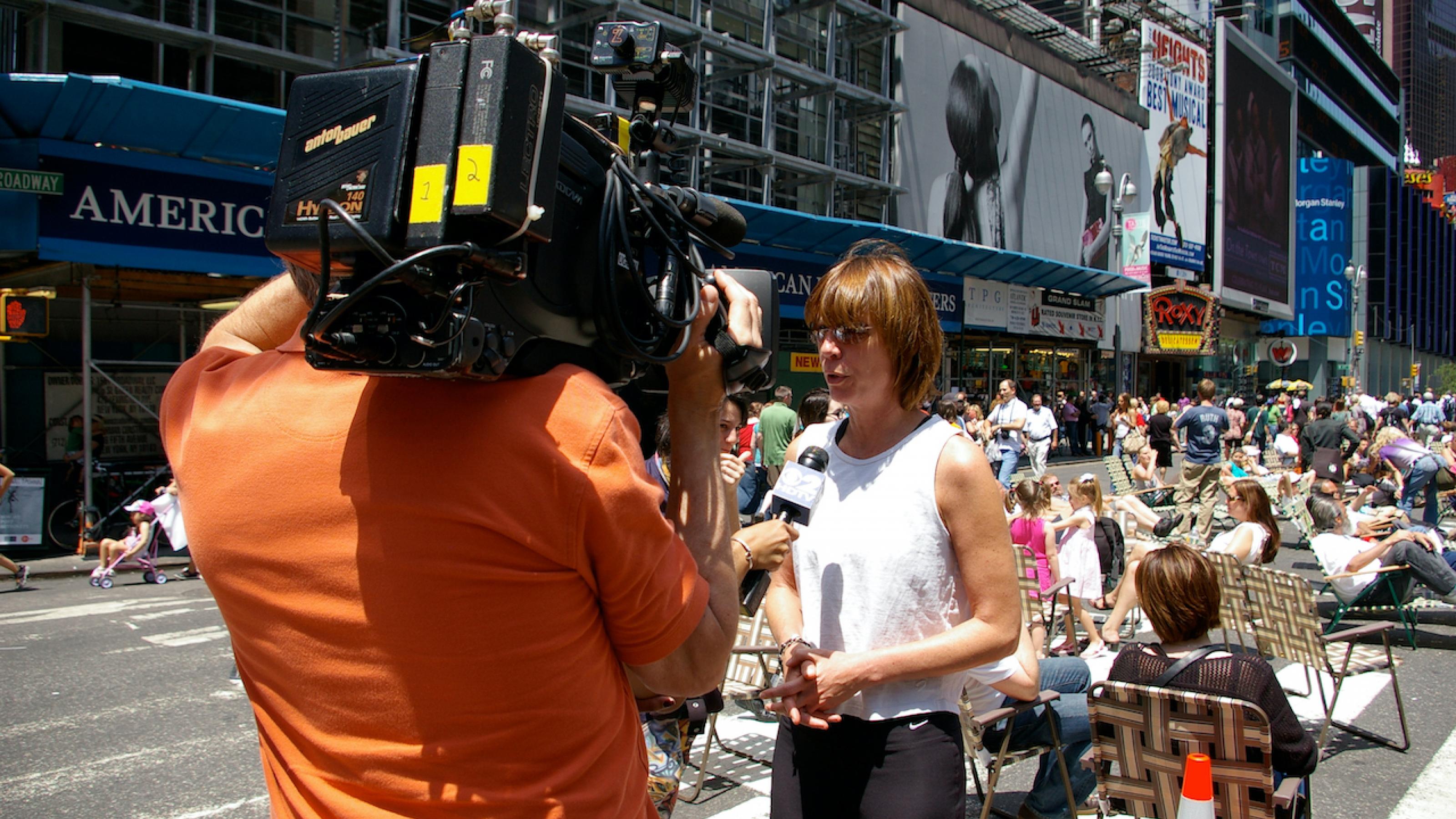 Janette Sadik-Khan steht auf dem für Autos gesperrten Abschnitt des Times-Square und wird gefilmt. Sie spricht in die Kamera. Im Hintergrund sieht man Menschen, die im Sonnenschein auf Stühlen sitzen