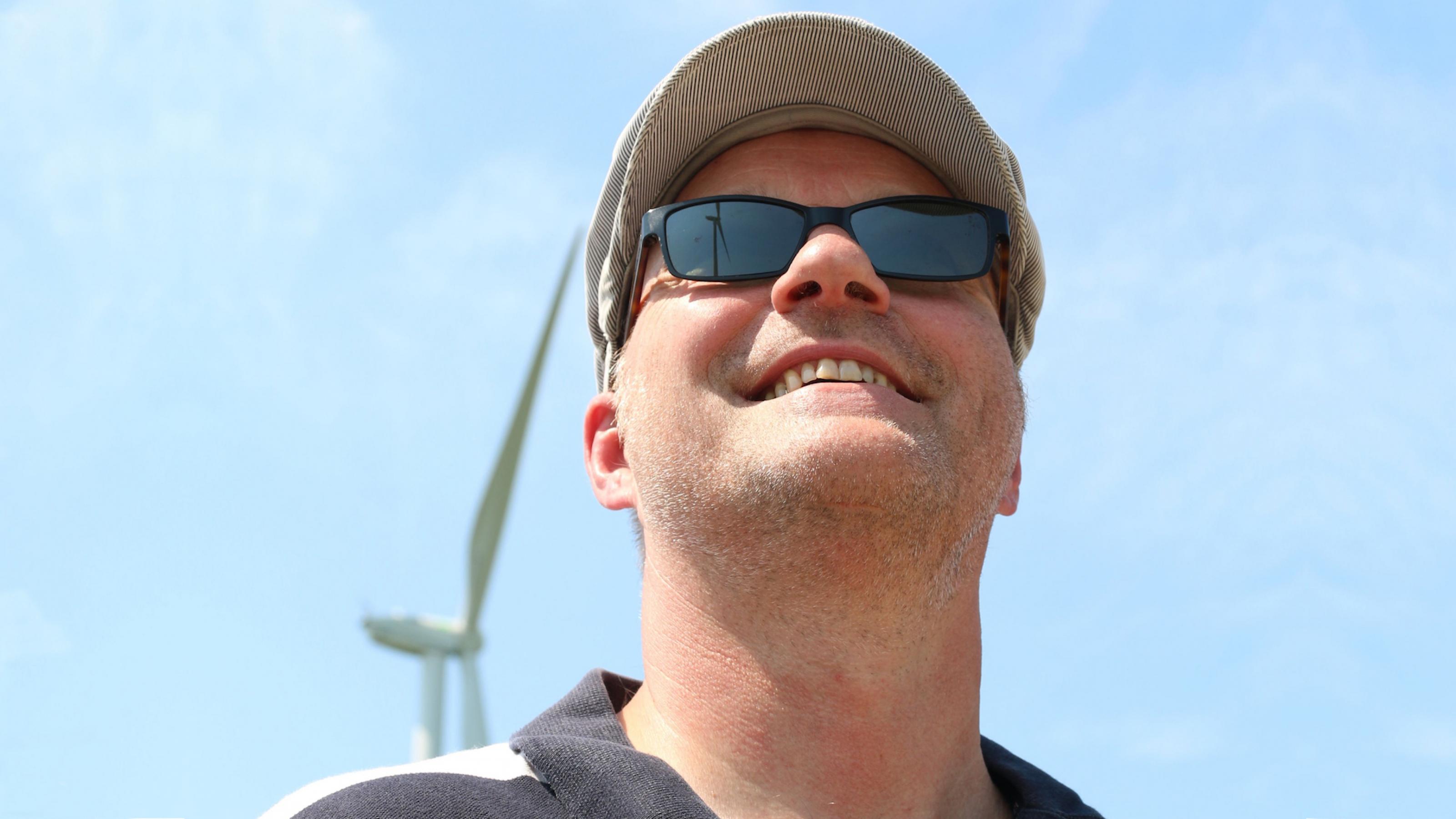 Ein Mann blickt nach oben in den Himmel. Über seine Schulter ist ein Windrad zu sehen, ein weiteres spiegelt sich in seiner Sonnenbrille. Jan Heidorn, der mehr als seit 25 Jahren Windräder in Hamburgs Südosten betreibt, in seinem Windpark im Stadtteil Neuengamme.