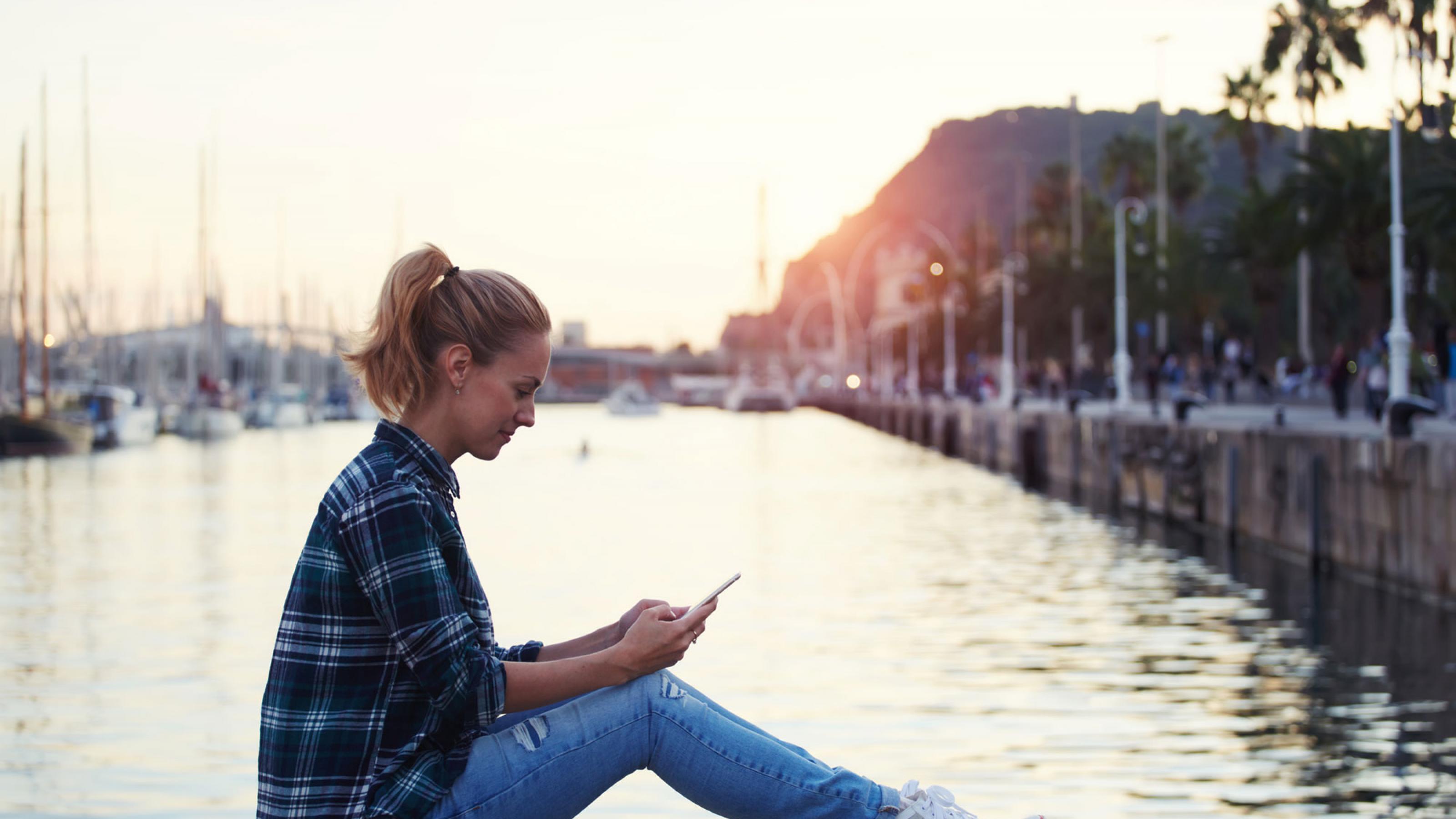 Eine junge Frau sitzt auf einer Kaimauer vor einem Hafenbecken und chattet lächelnd per Smartphone