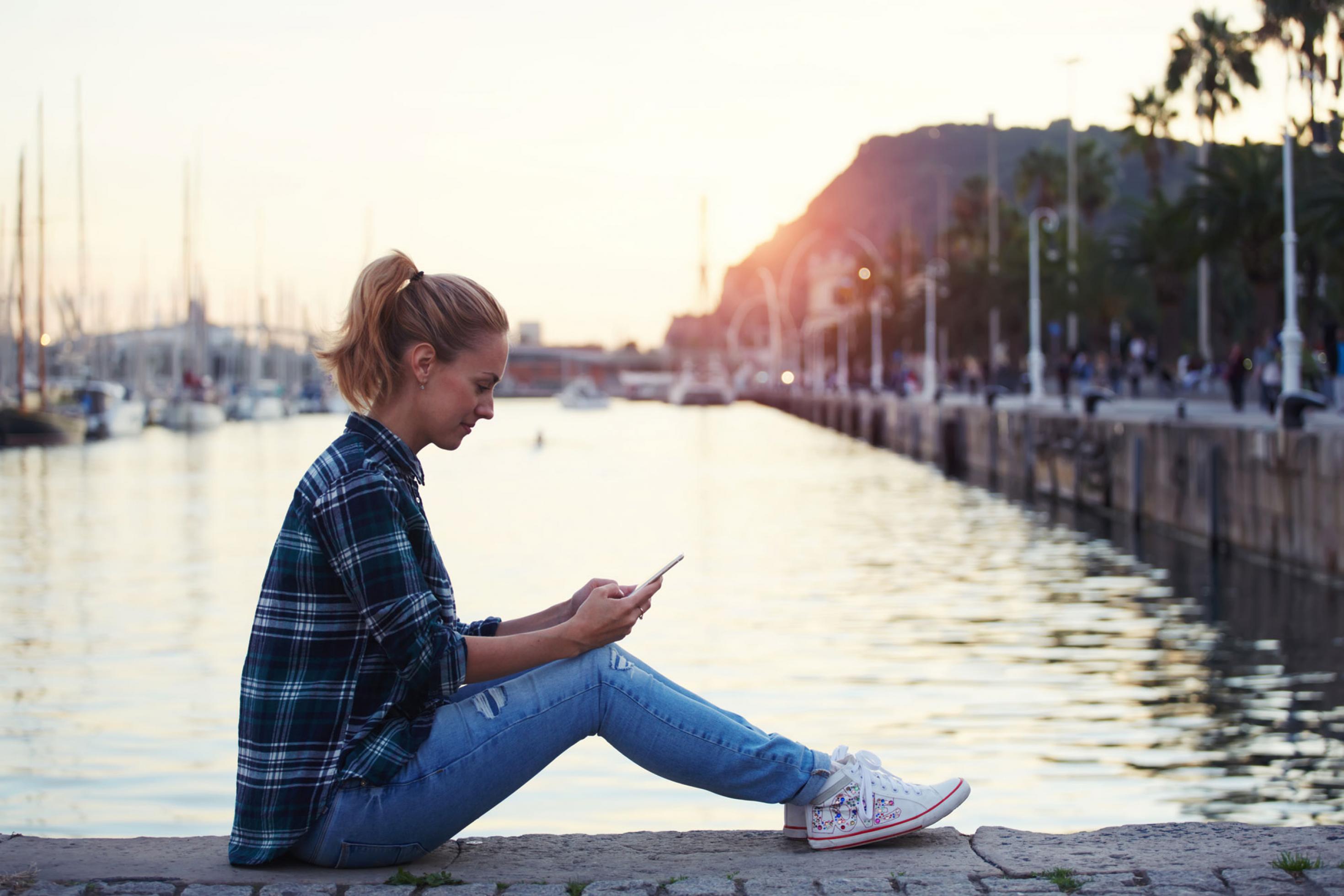Eine junge Frau sitzt auf einer Kaimauer vor einem Hafenbecken und chattet lächelnd per Smartphone