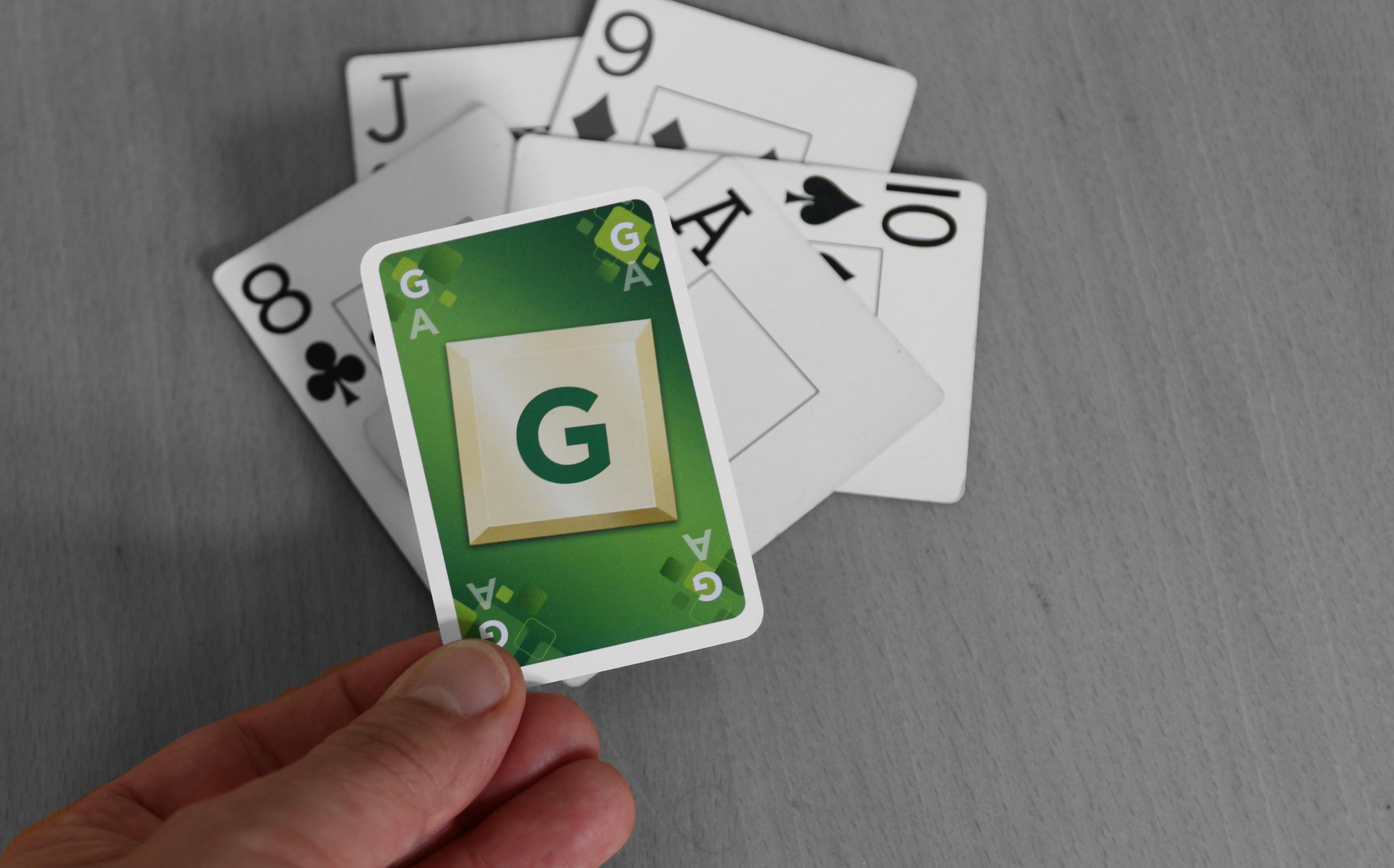 Auf einen Tisch liegen fünf Spielkarten. Eine Hand legt eine sechste Karte darauf. Sie gehört aber nicht zu einem normalen Kartenspiel, sondern ist leuchtend grün und zeigt ein großes G. Die Tischoberfläche und die Karten darauf sind hingegen in schwarz-weiß gehalten. – Ein grüner Trumpf für einen grauen Stich.