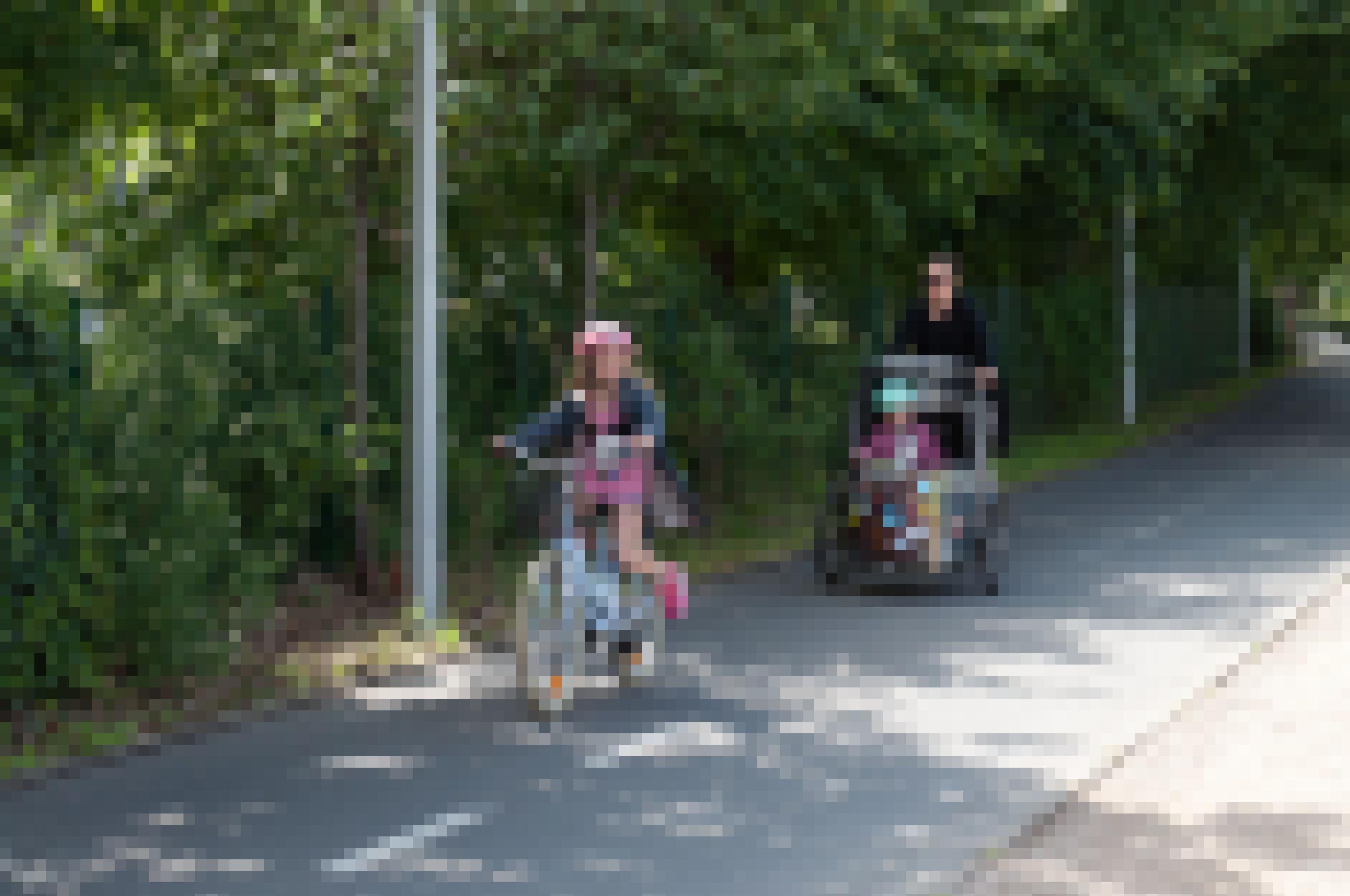 Ein kleines Mädchen fährt Fahrrad auf einem breiten Radweg. Einige Meter hinter ihr fährt die Mutter.