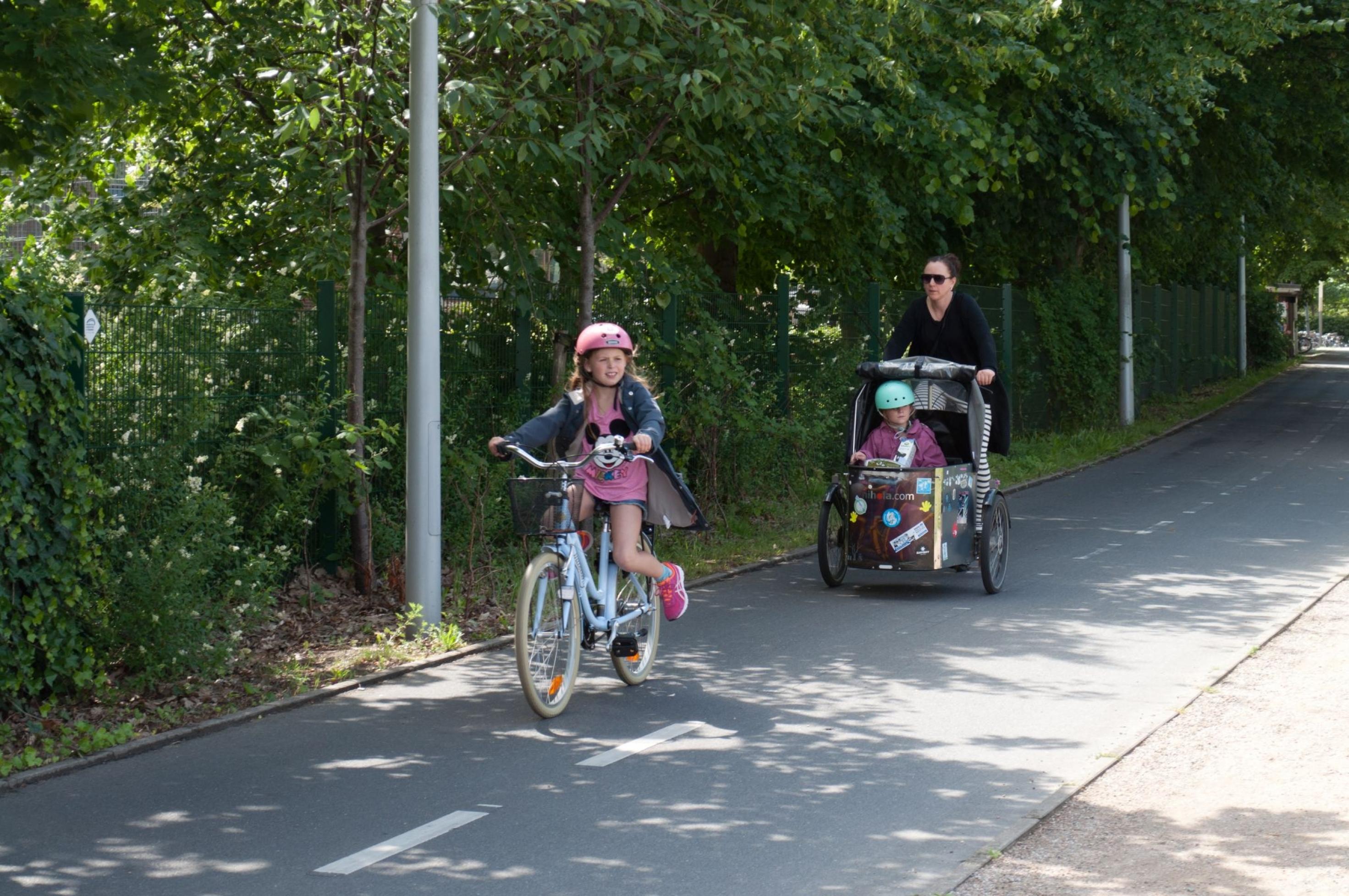 Ein kleines Mädchen fährt Fahrrad auf einem breiten Radweg. Einige Meter hinter ihr fährt die Mutter.