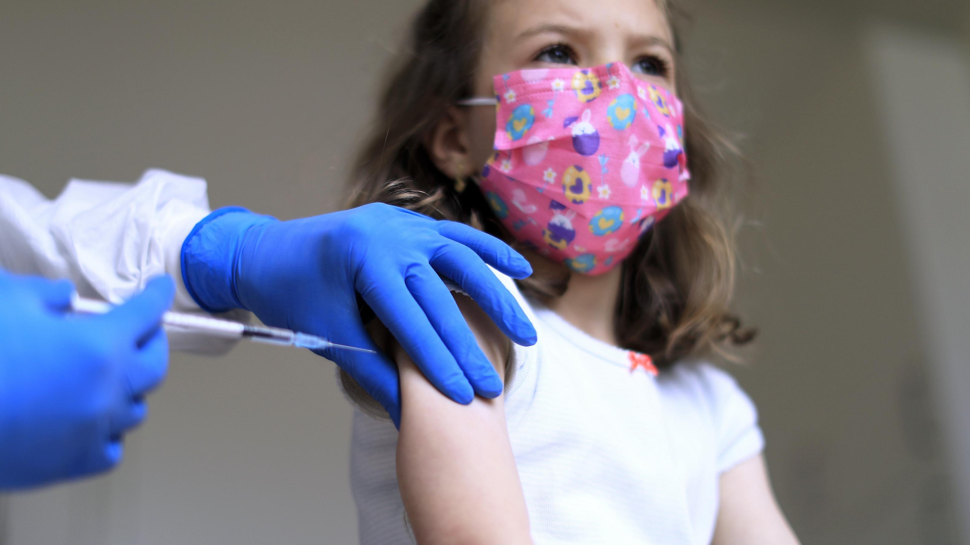 Ein Arzt führt eine Spritze mit Impfstoff an den rechten Arm eines Mädchens, das eine rosafarbene Maske trägt und in die Ferne schaut.