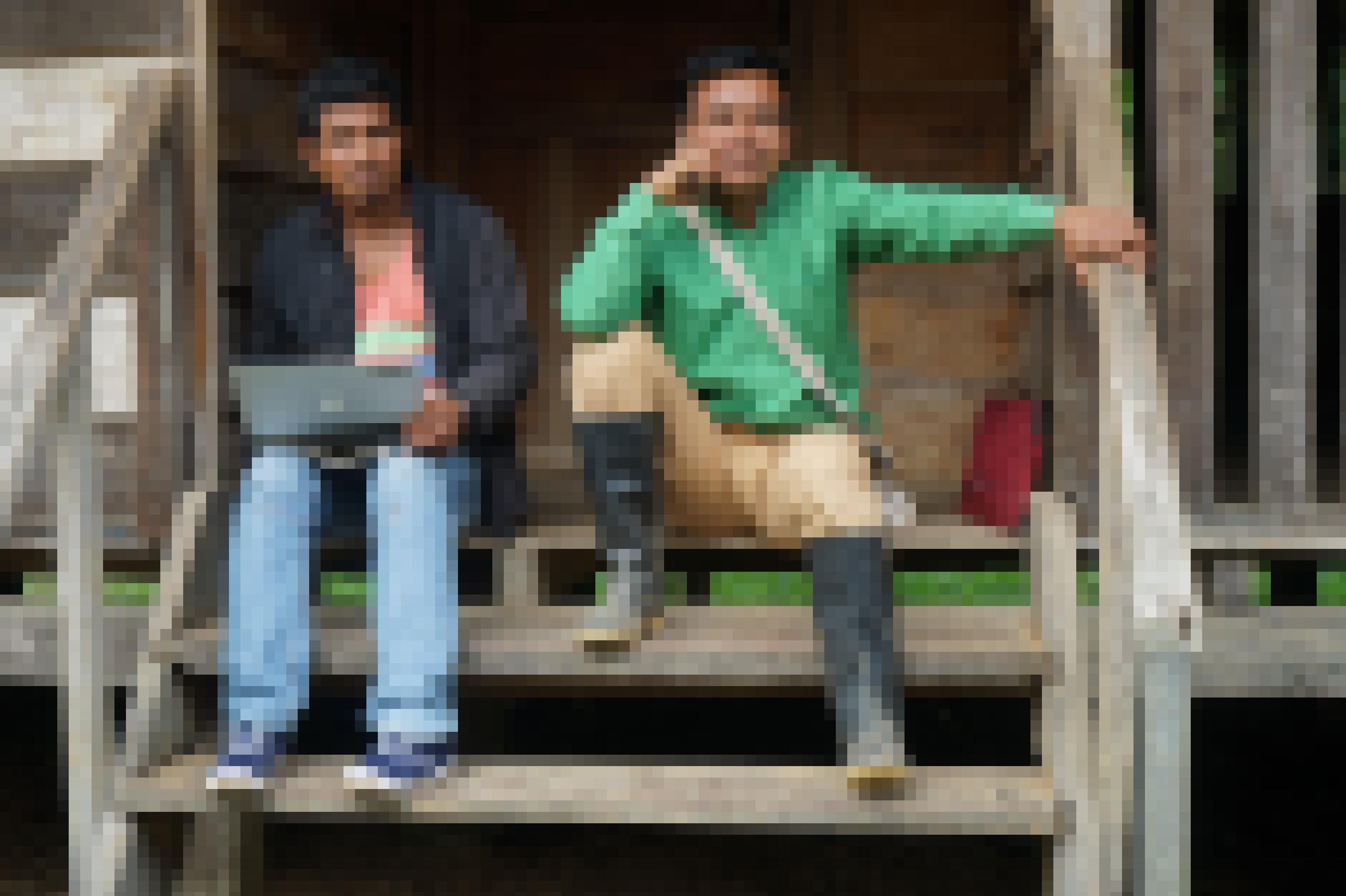 Zwei junge Männer, einer in Gummistiefeln, sitzen mit Handy am Ohr und Laptop auf einer Holztreppe im Dschungel.