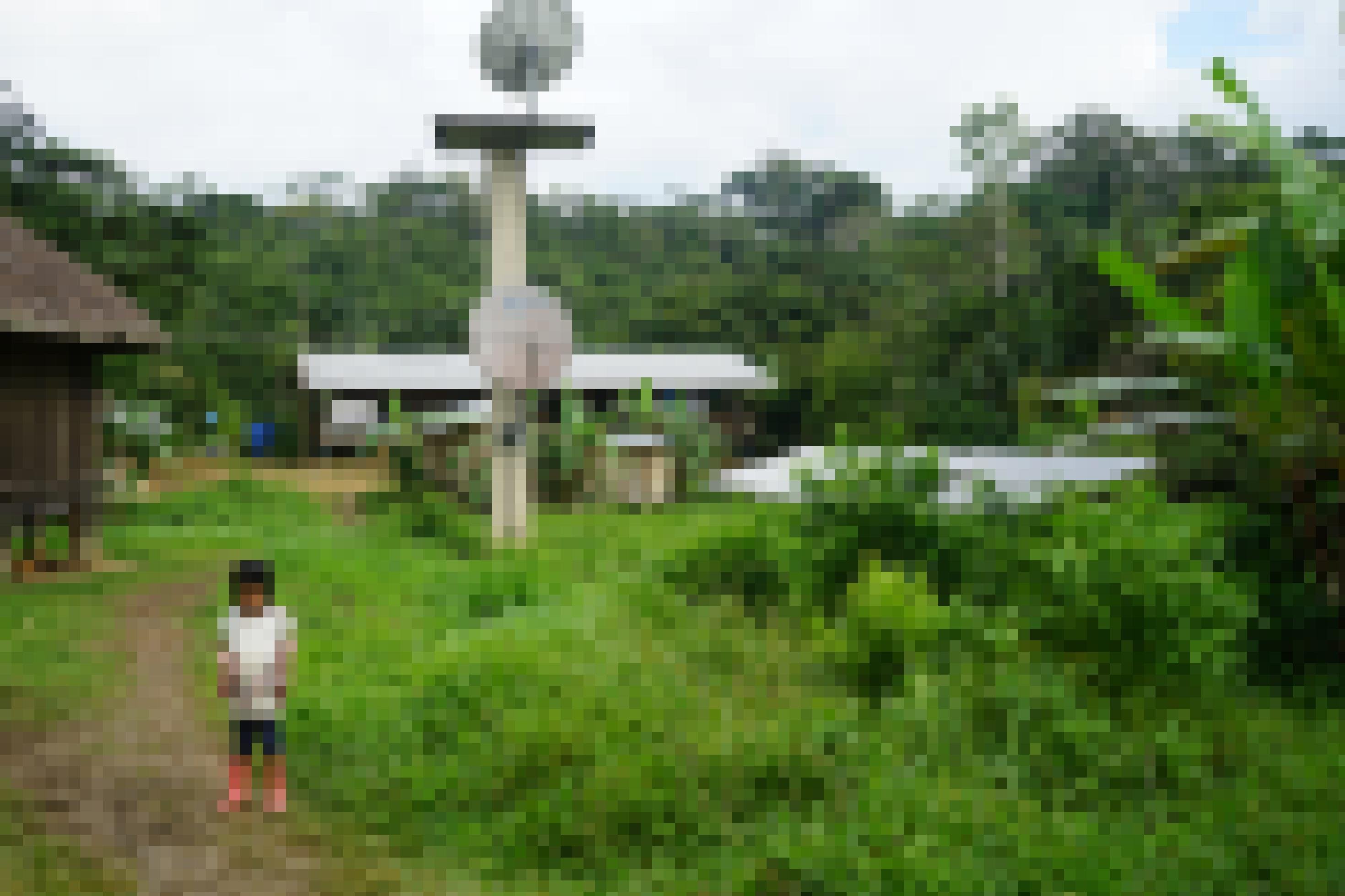 Solarpanel und Satellitenschüsseln neben Holzhütten, dahinter Urwald, im Vordergrund läuft ein indigenes Kind auf einem Pfad.