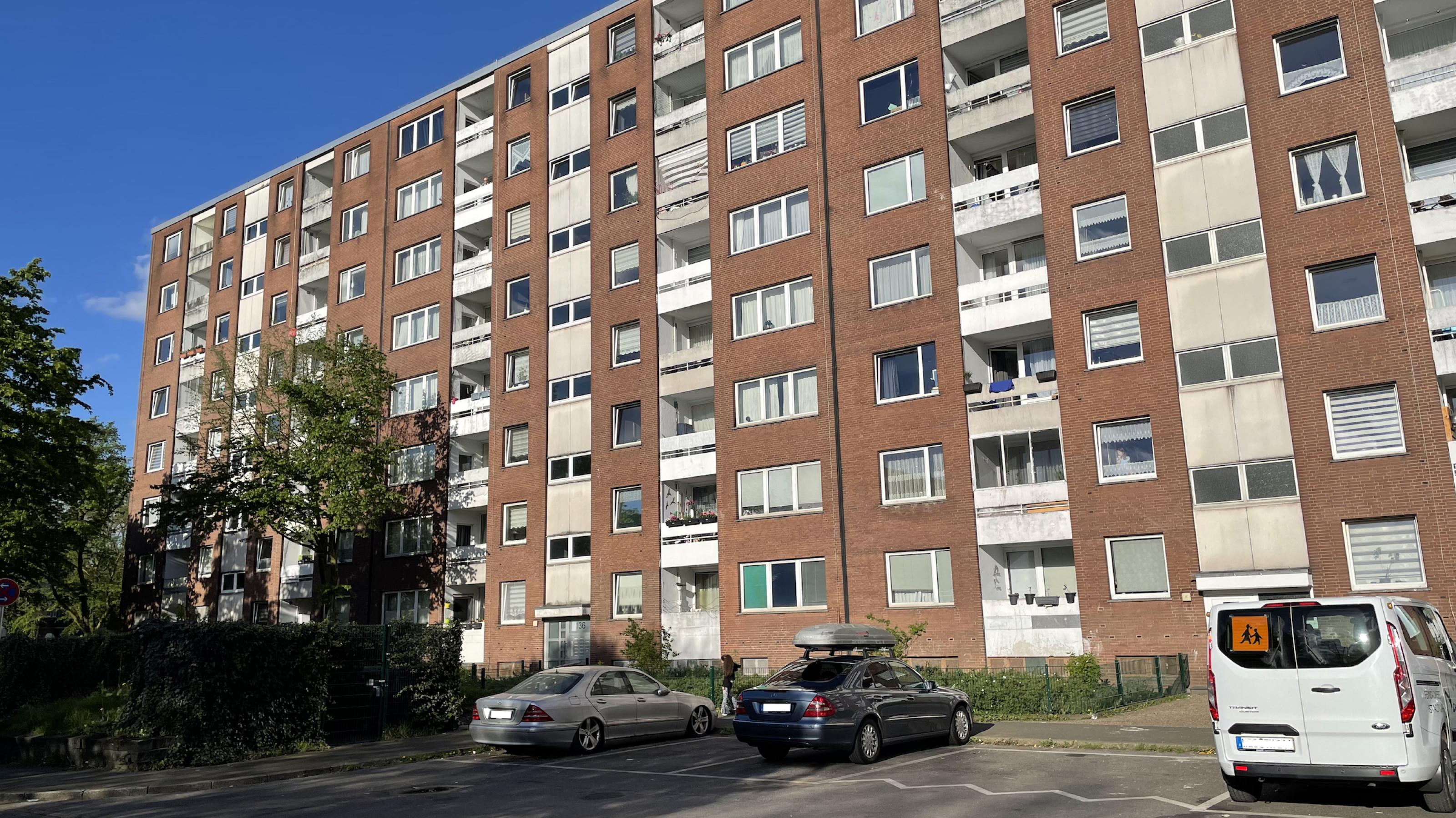 Siebenstöckiger Block von Mietwohnungen einfachen Standards mit Backsteinfassade und Flachdach. Vor dem Haus stehen auf einem Parkplatz drei Autos.