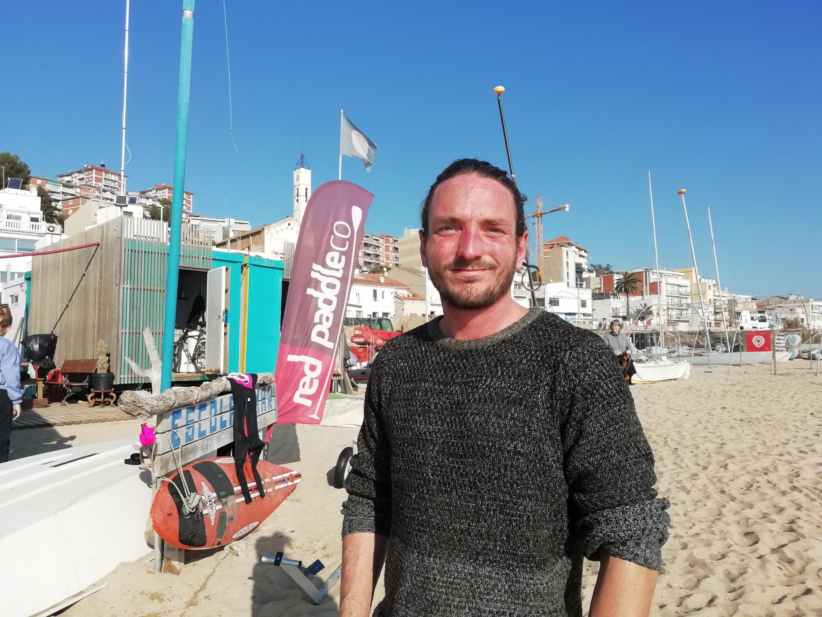 Der Leiter der Segelschule Escola de Mar steht an einem sonnigen Wintertag am Strand, im Hintergrund ist die Segelschule und die Kulisse der Ortschaft Montgat zu sehen.