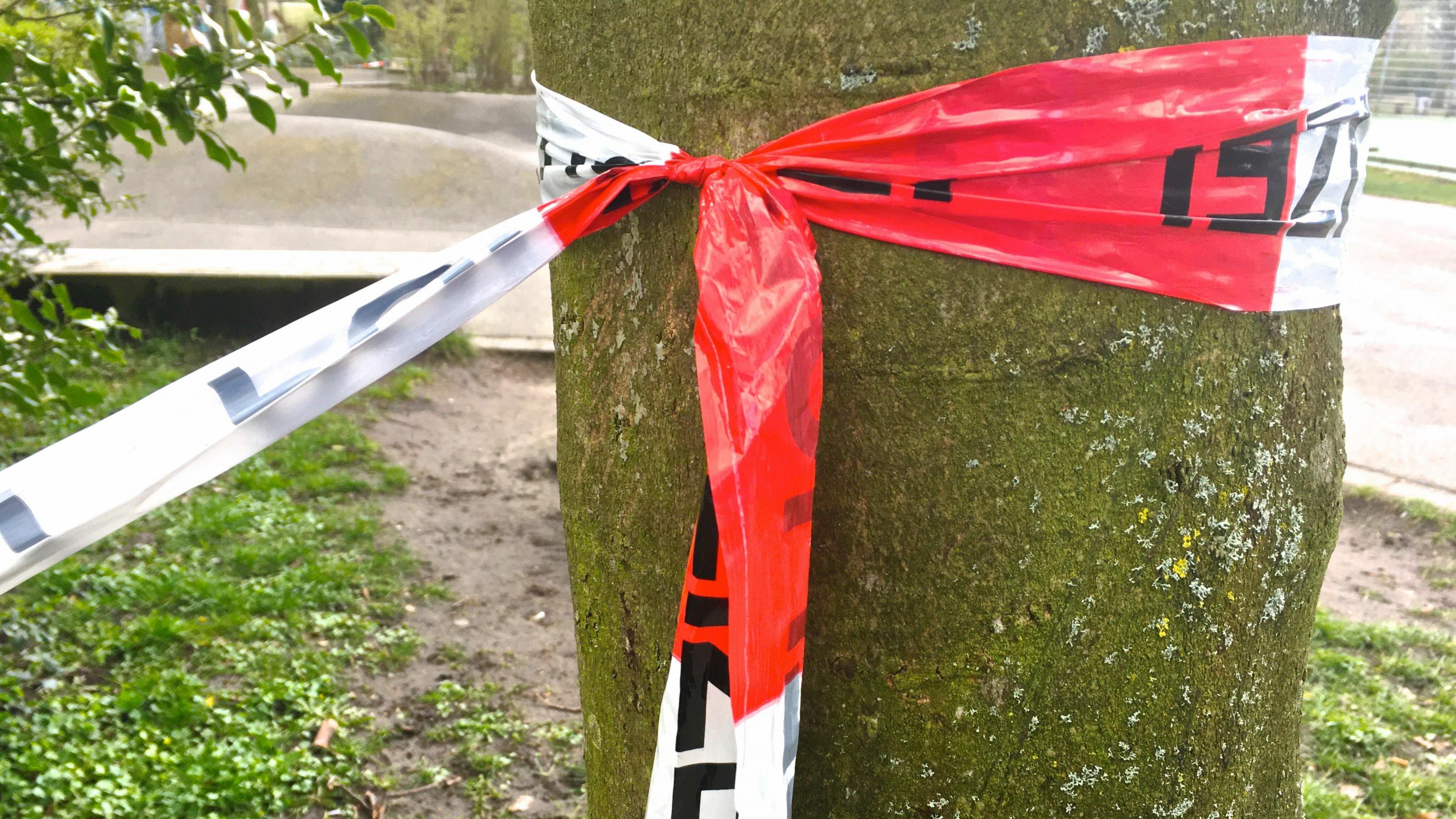 Ein rot-weißes Flatterband ist um einen Baumstamm geschlungen und dort verknotet. Er dient als Haltepunkt für die Absperrung eines Spielplatzes.