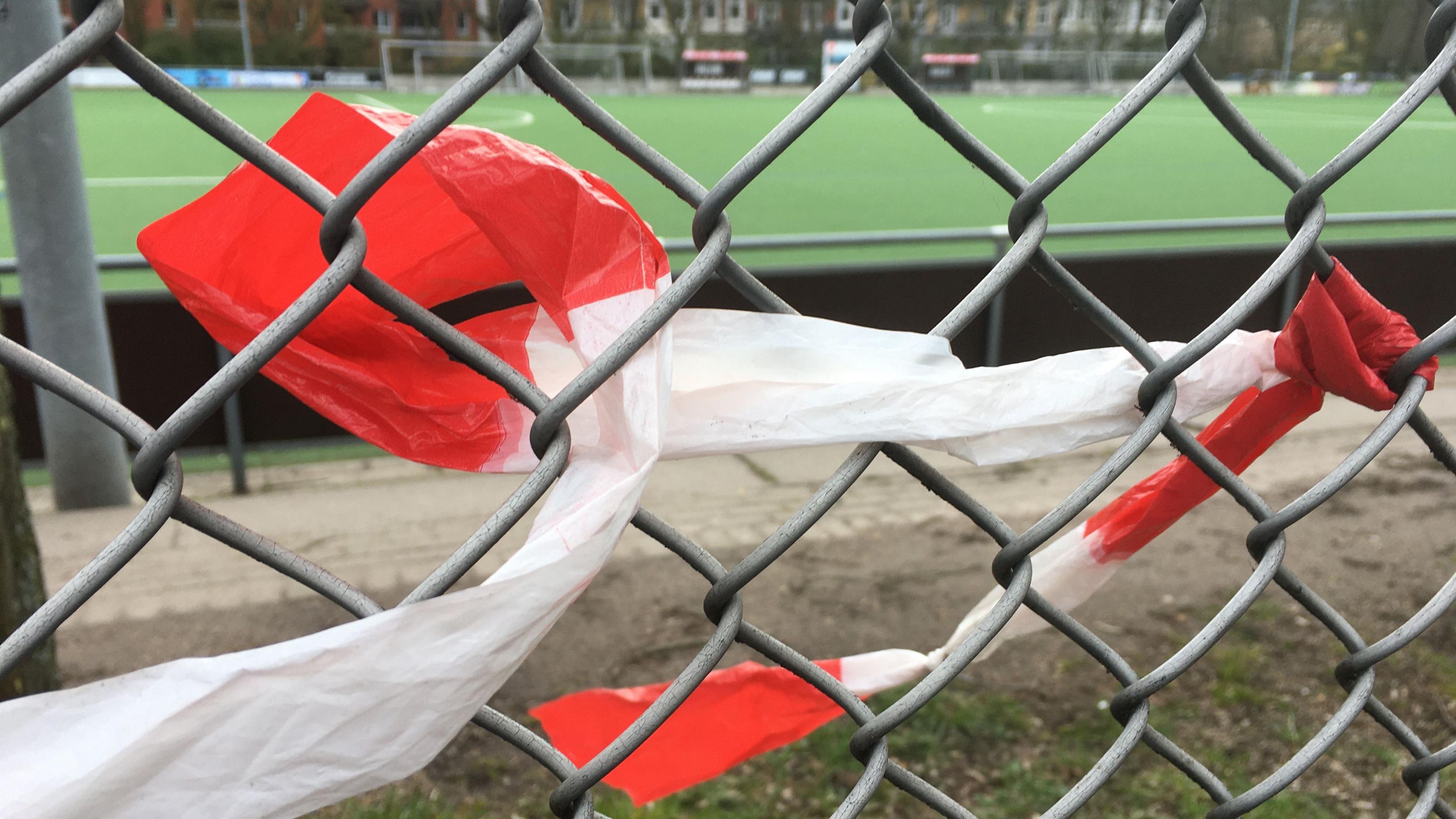 Ein rot-weißes Flatterband ist an einem Draht eines Maschendrahtzauns festgeknotet und windet sich von da nach links locker durch das Geflecht. Im Hintergrund ist ein Fußballplatz mit hellgrünen Kunststoff-Belag zu sehen.