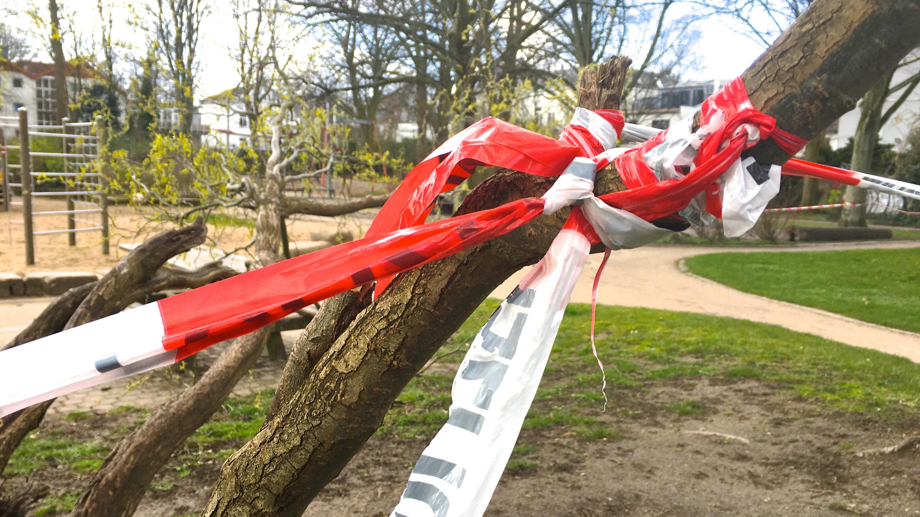 Ein knorriger Ast, um den ein rot-weißes Absperrband mit dem Aufdruck „Polizei“ gewickelt und geknotet ist. Es sperrt den Zugang zum Spielplatz in einem Hamburger Park ab.