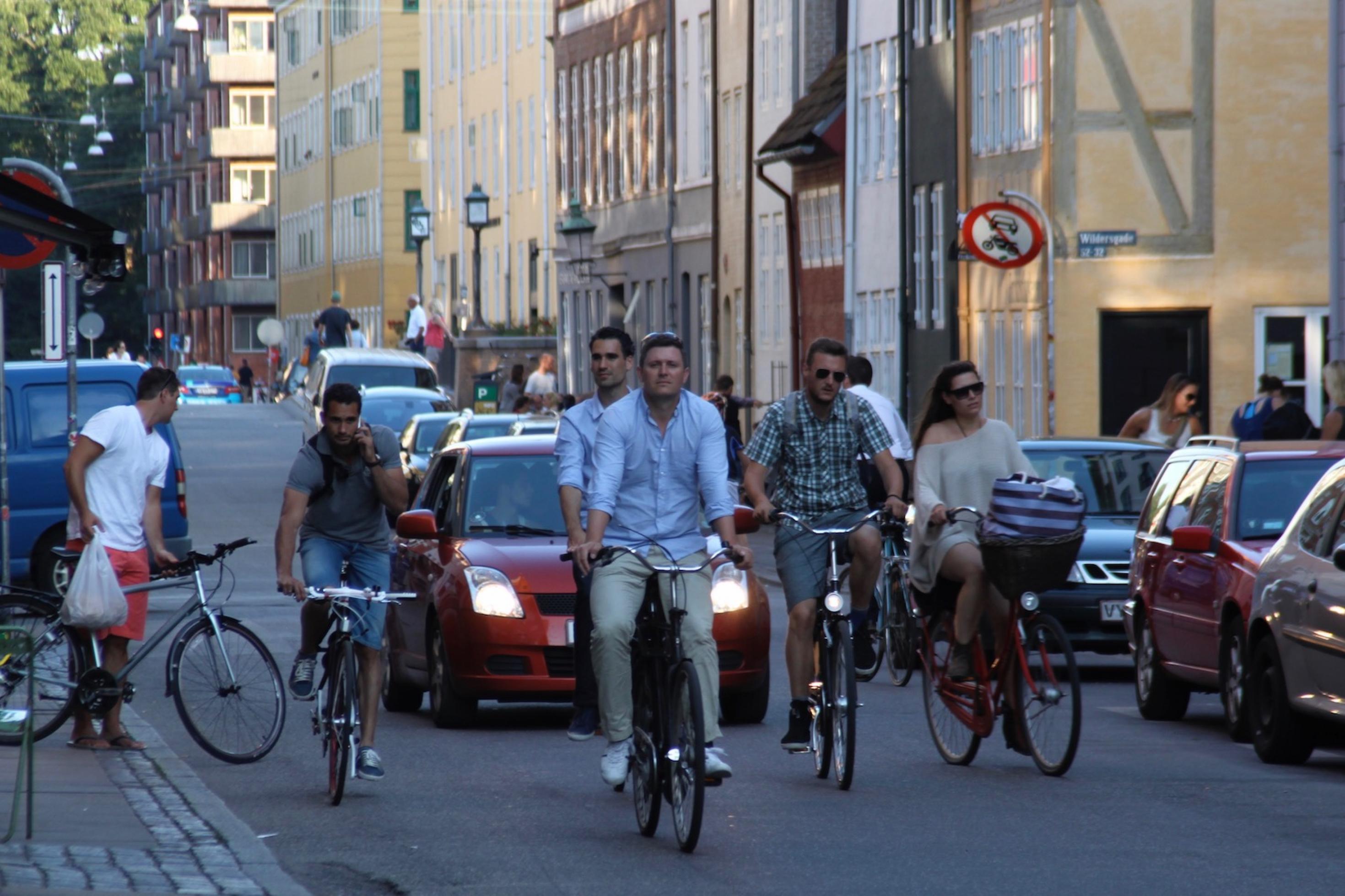 Auf einer kleinen Straße fahren viele Radfahrer. Hinter ihnen stauen sich einige Autos.