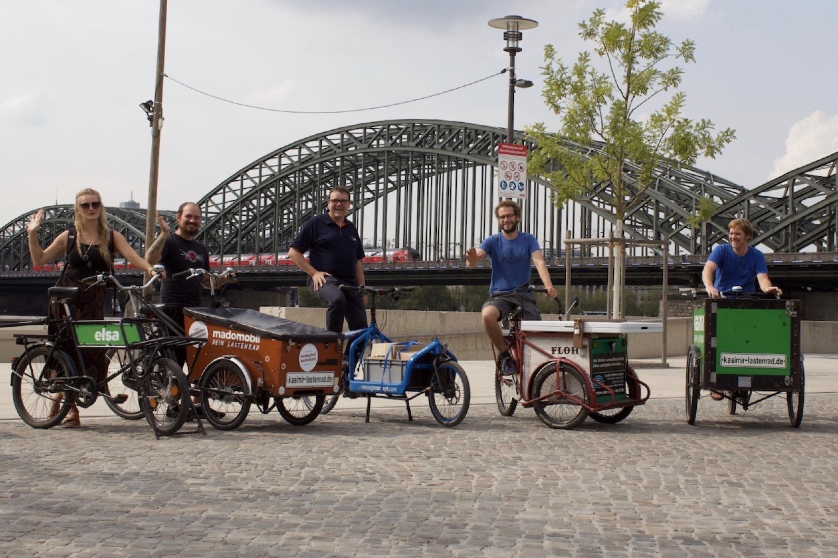 Fünf Fahrer und Fahrerinnen posieren auf unterschiedlichen Transporträdern mit zwei und drei Rädern auf einem freien Platz. Im Hintergrund sieht man die Silhouette der Hohenzollern Brücke.