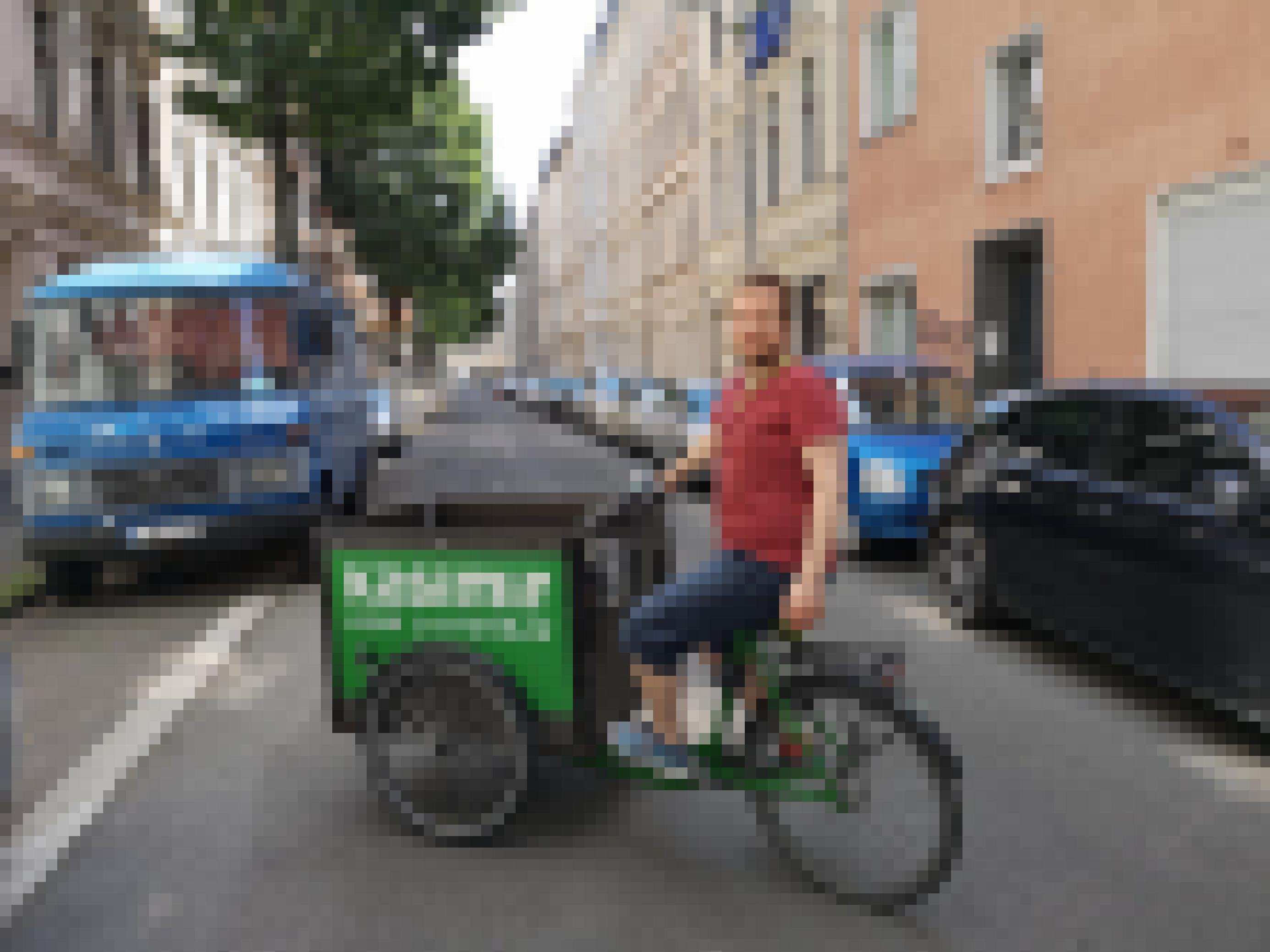 Ein Mann mit kurzen braunen Haaren und Bart sitzt auf einem Lastenrad mit grüner Box. Er trägt ein rotes T-Shirt und blaue, halblange Jeans.