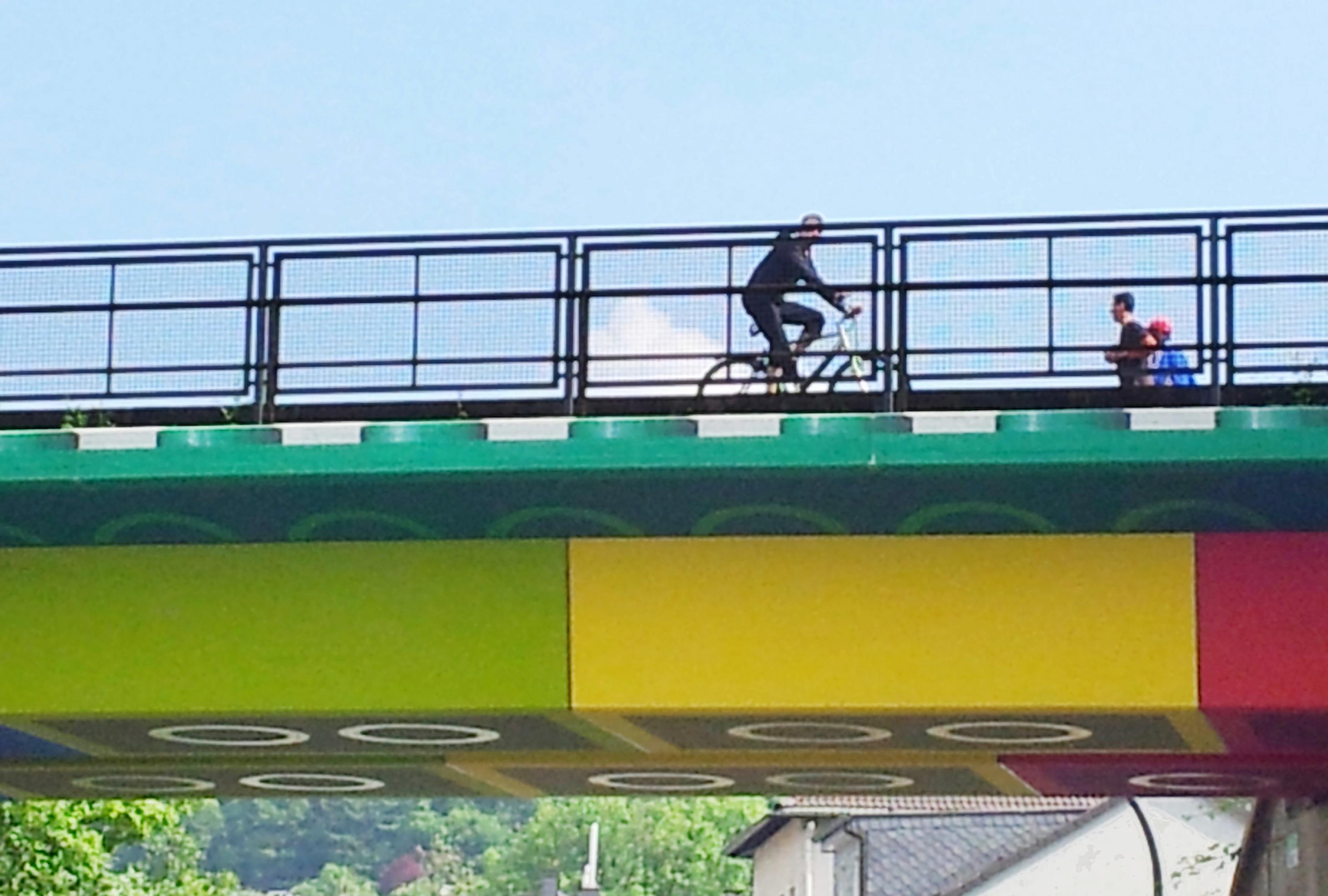 Ein Ausschnitt der Lego-Brücke, auf der ein Radfahrer und zwei Fußgänger hinter dem Geländer zu sehen sind. Der Himmel ist blau.