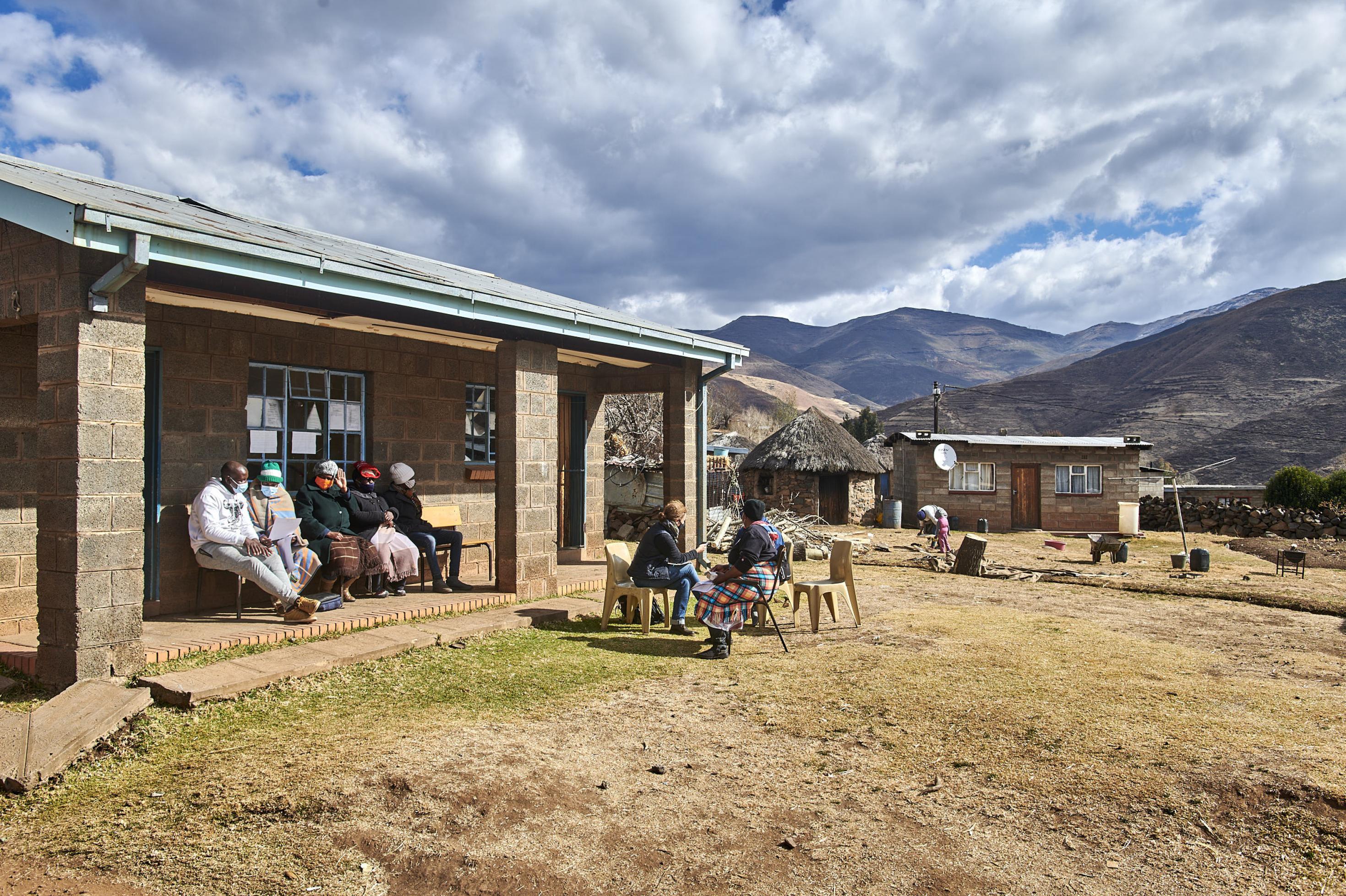 Vor einem kleinen Haus sitzen Bürger und Bürgerinnen, traditionelle Rundhütten und die Berge im Hintergrund